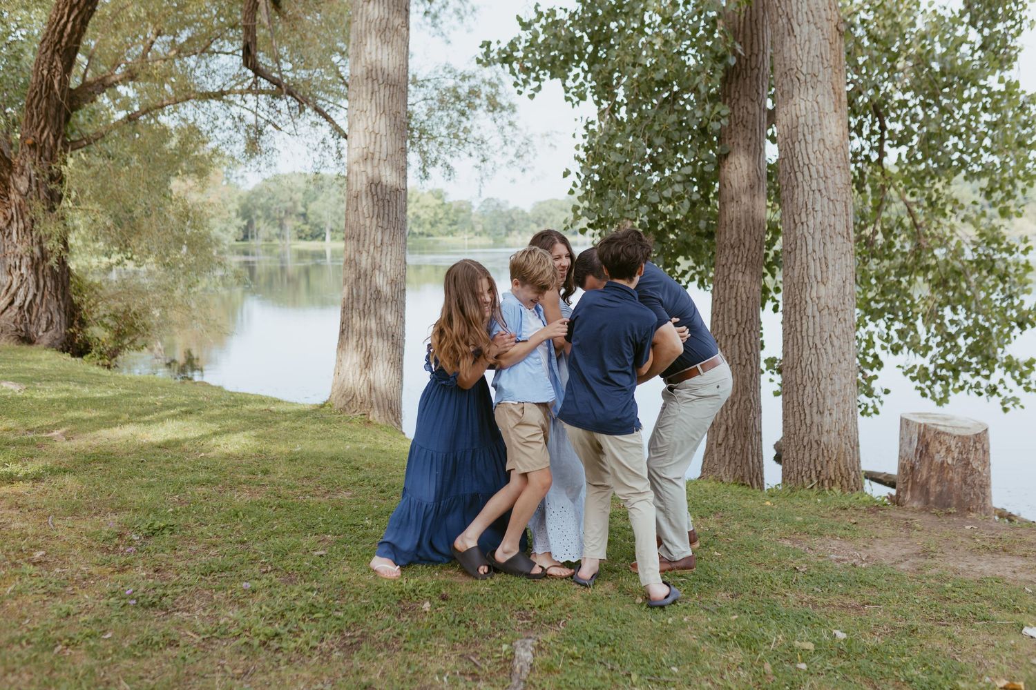 Family of five sharing a warm embrace near a serene lake surrounded by tall trees during an outdoor photo session.