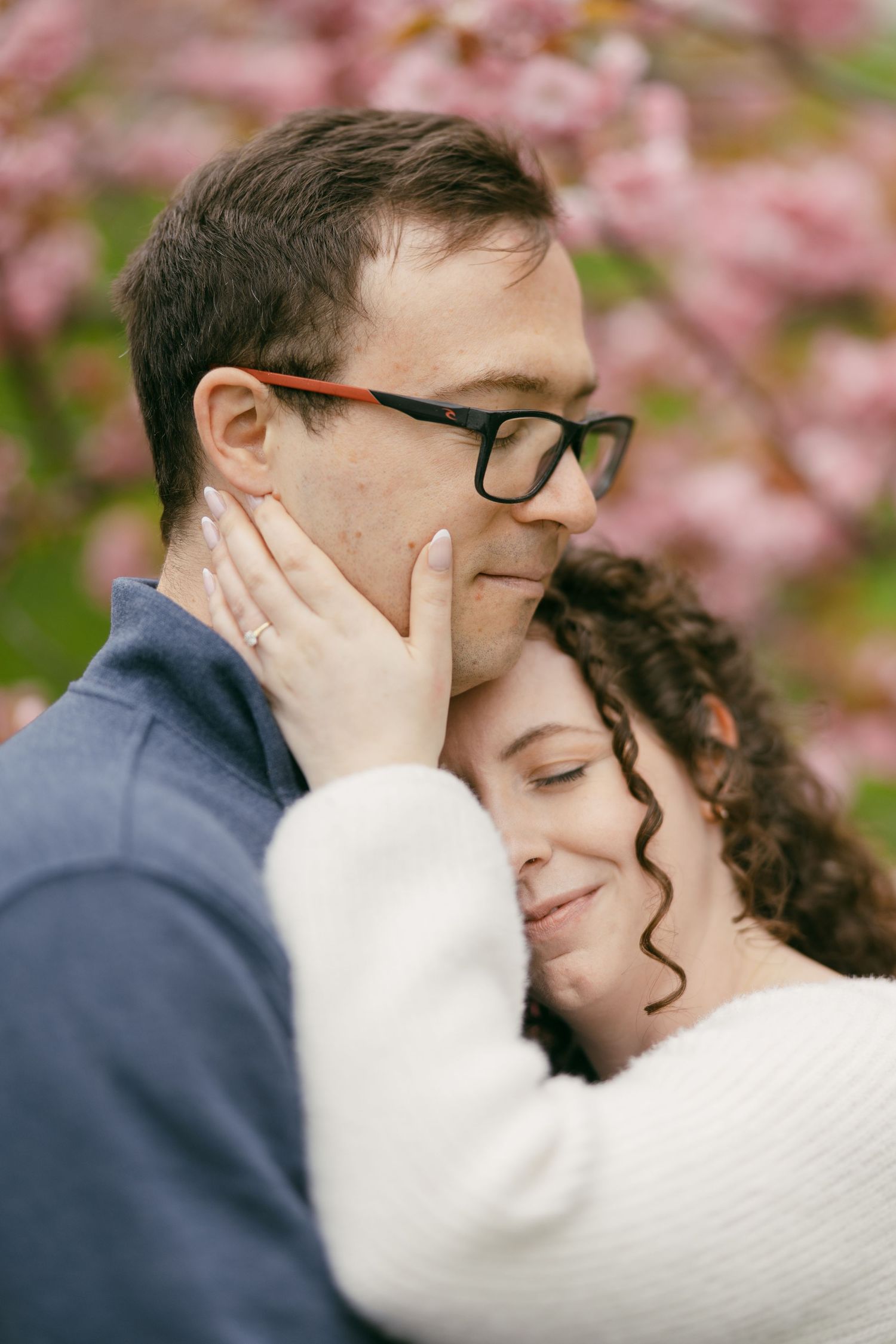 A couple embraces tenderly near pink cherry blossoms, the man wearing glasses as the woman rests her head on his cheek.