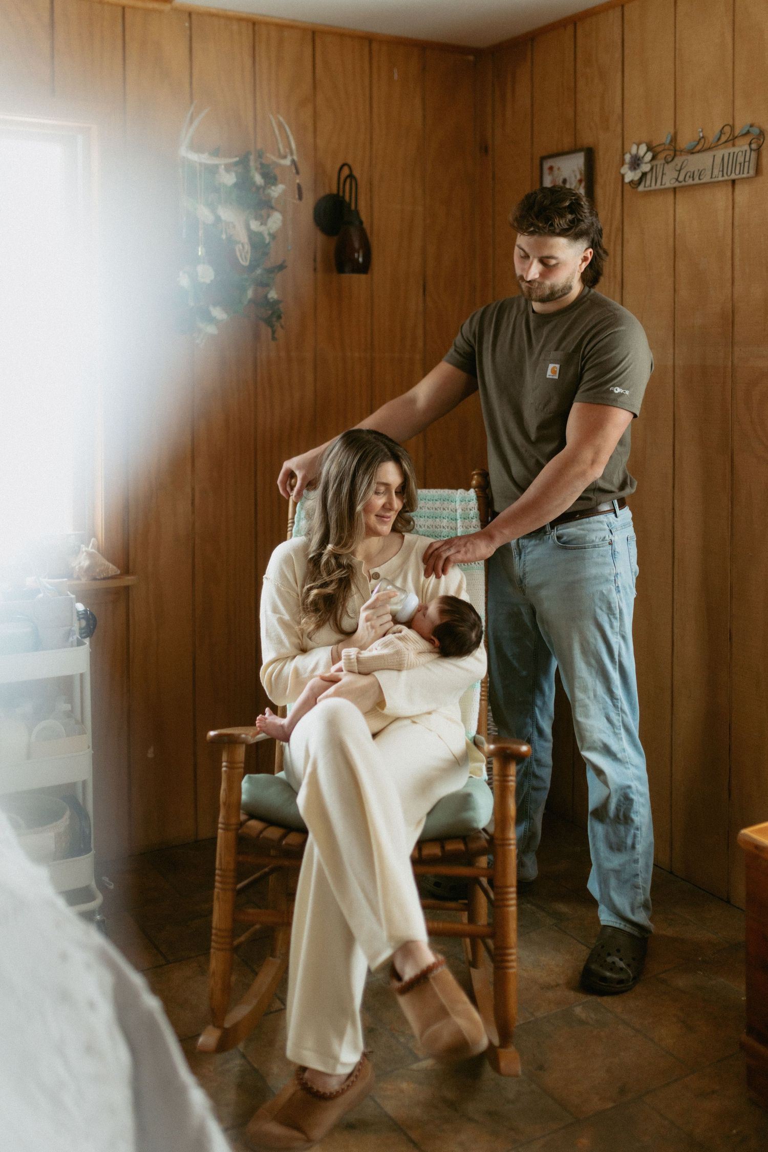 New parents share a tender moment, mother seated in rocking chair holding newborn while father stands beside them.