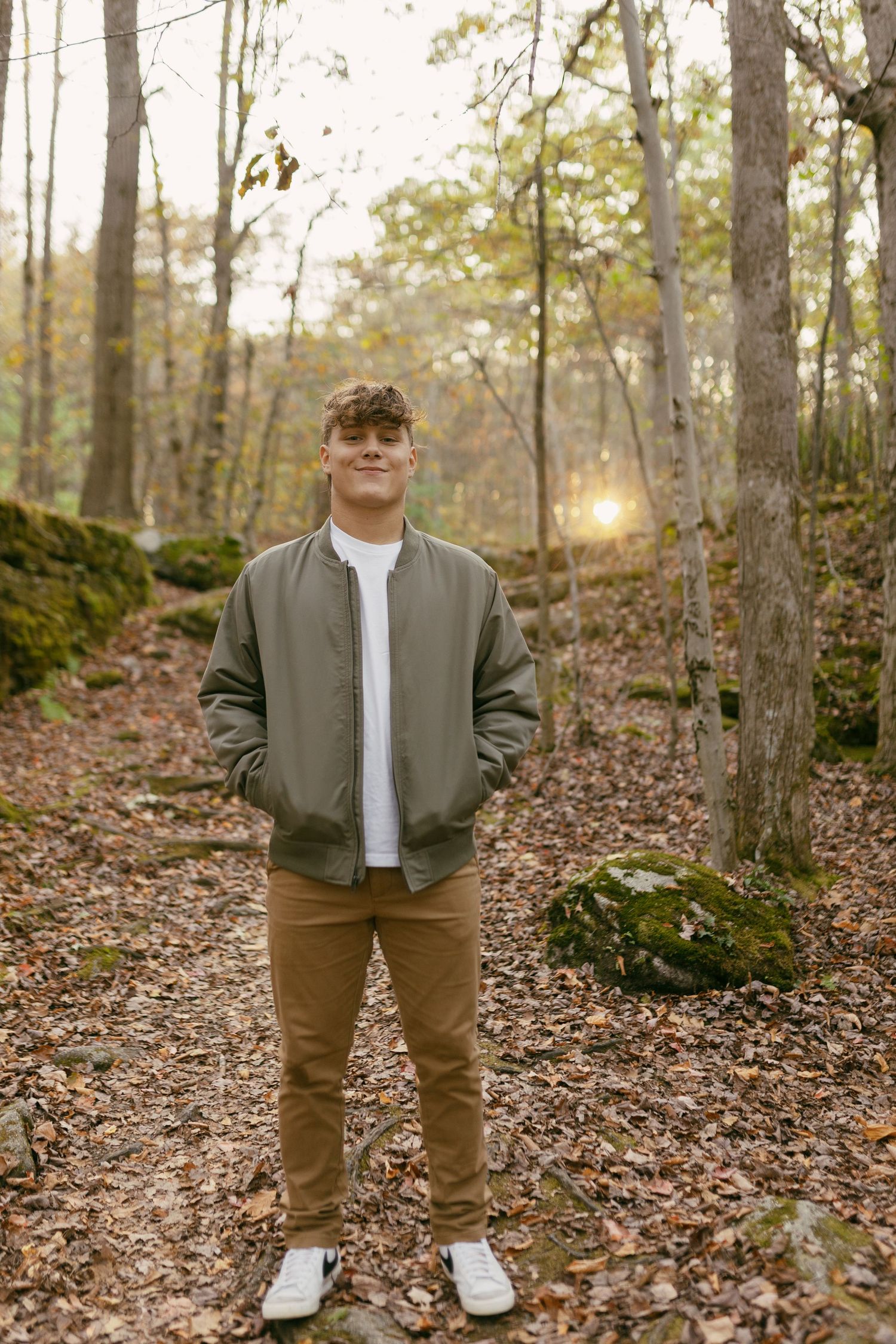 Young man in olive bomber jacket and tan pants stands smiling on a leaf-covered forest path at sunset.