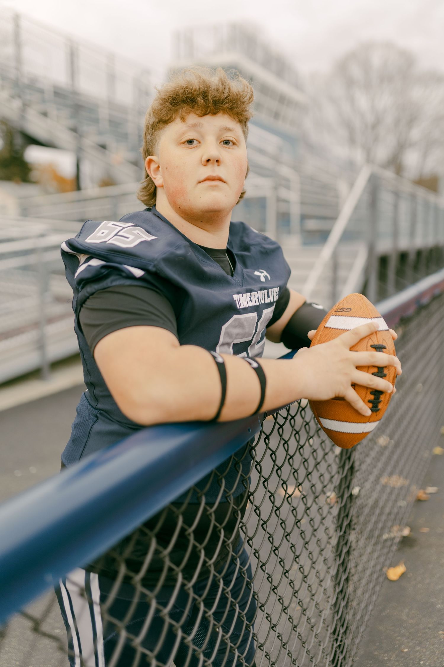 Young football player in navy jersey holding a football while leaning on a chain-link fence at a stadium.