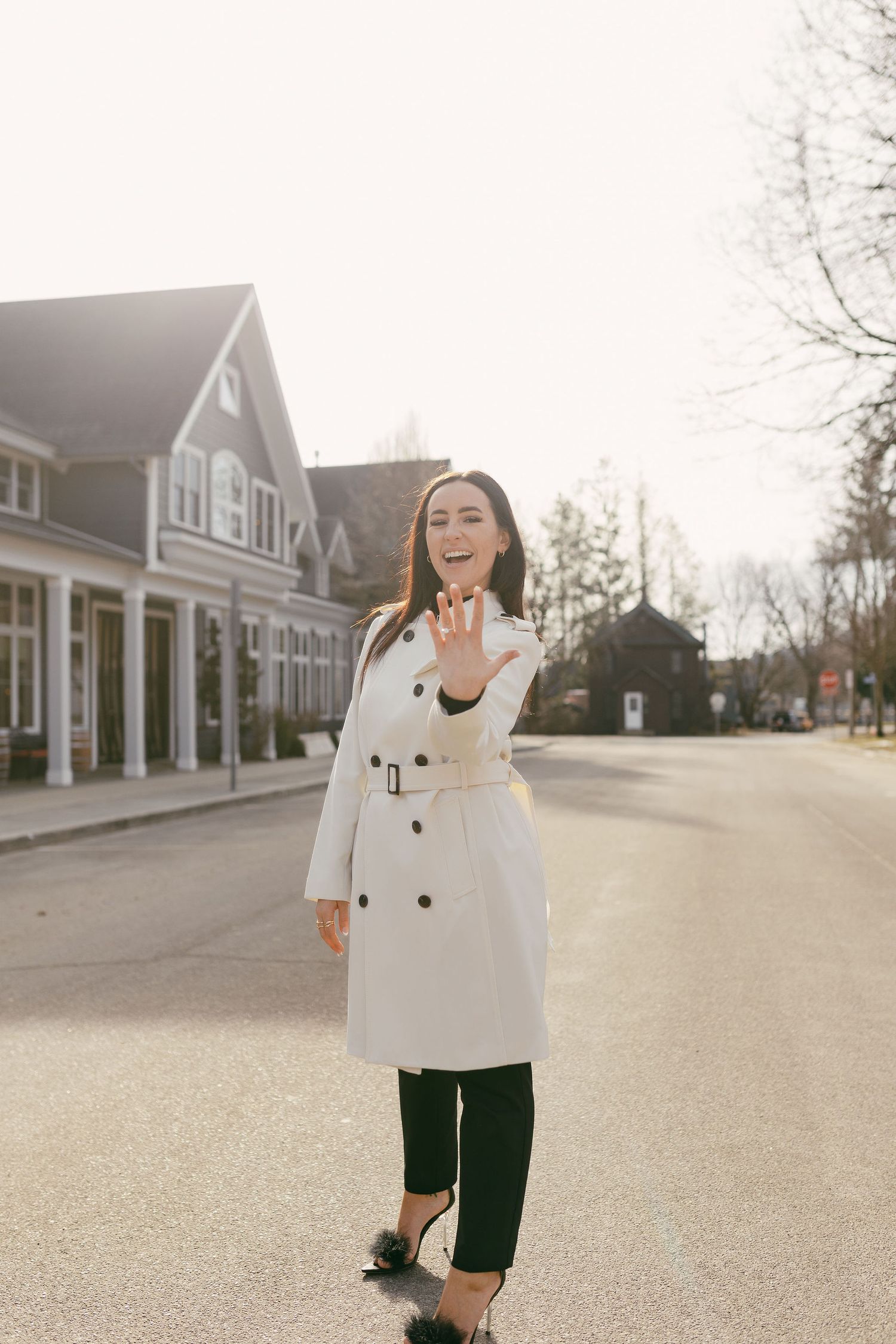 Smiling woman in white trench coat showing engagement ring on sunny suburban street.