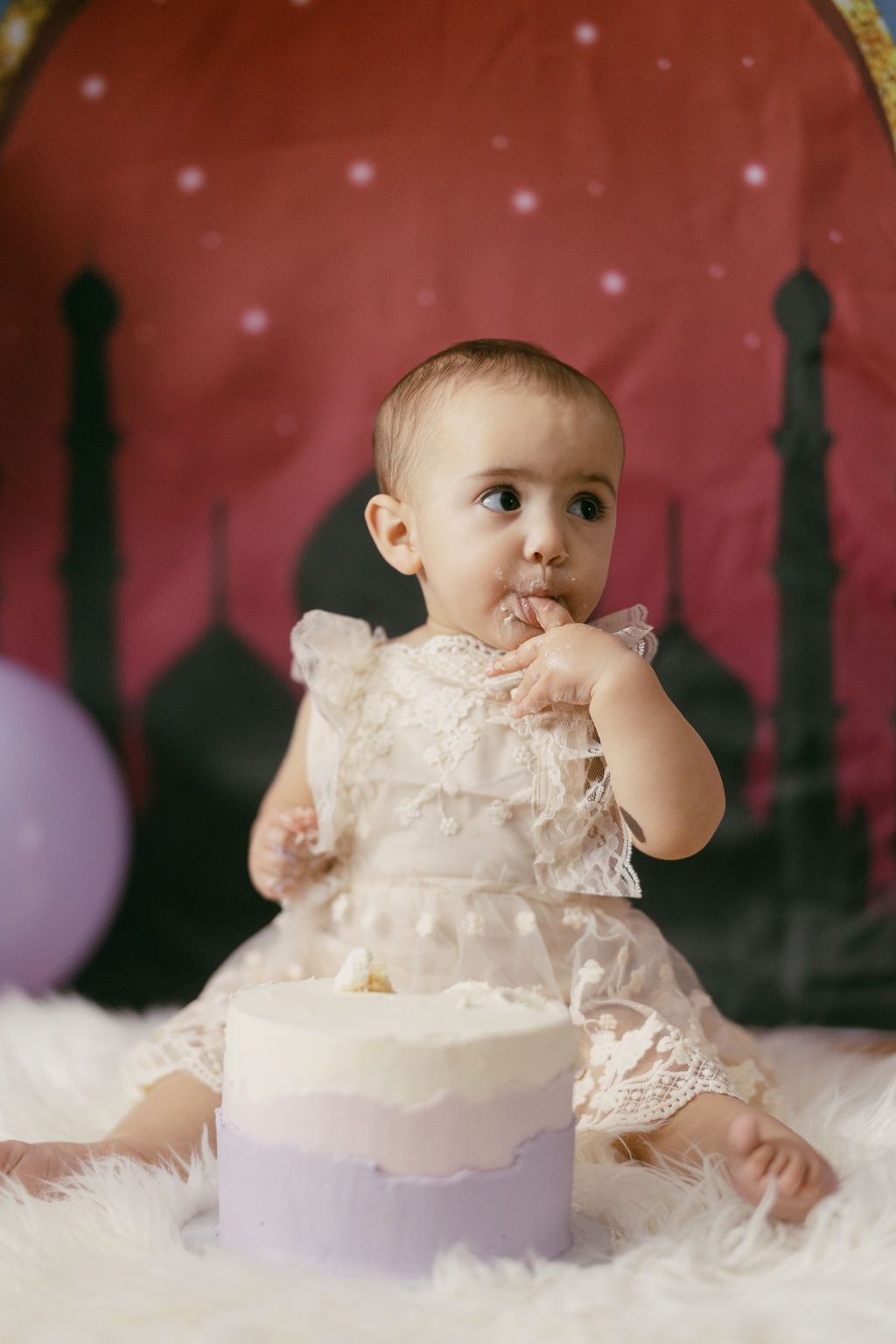 Baby in white lace dress photographed against red backdrop with twinkling lights.