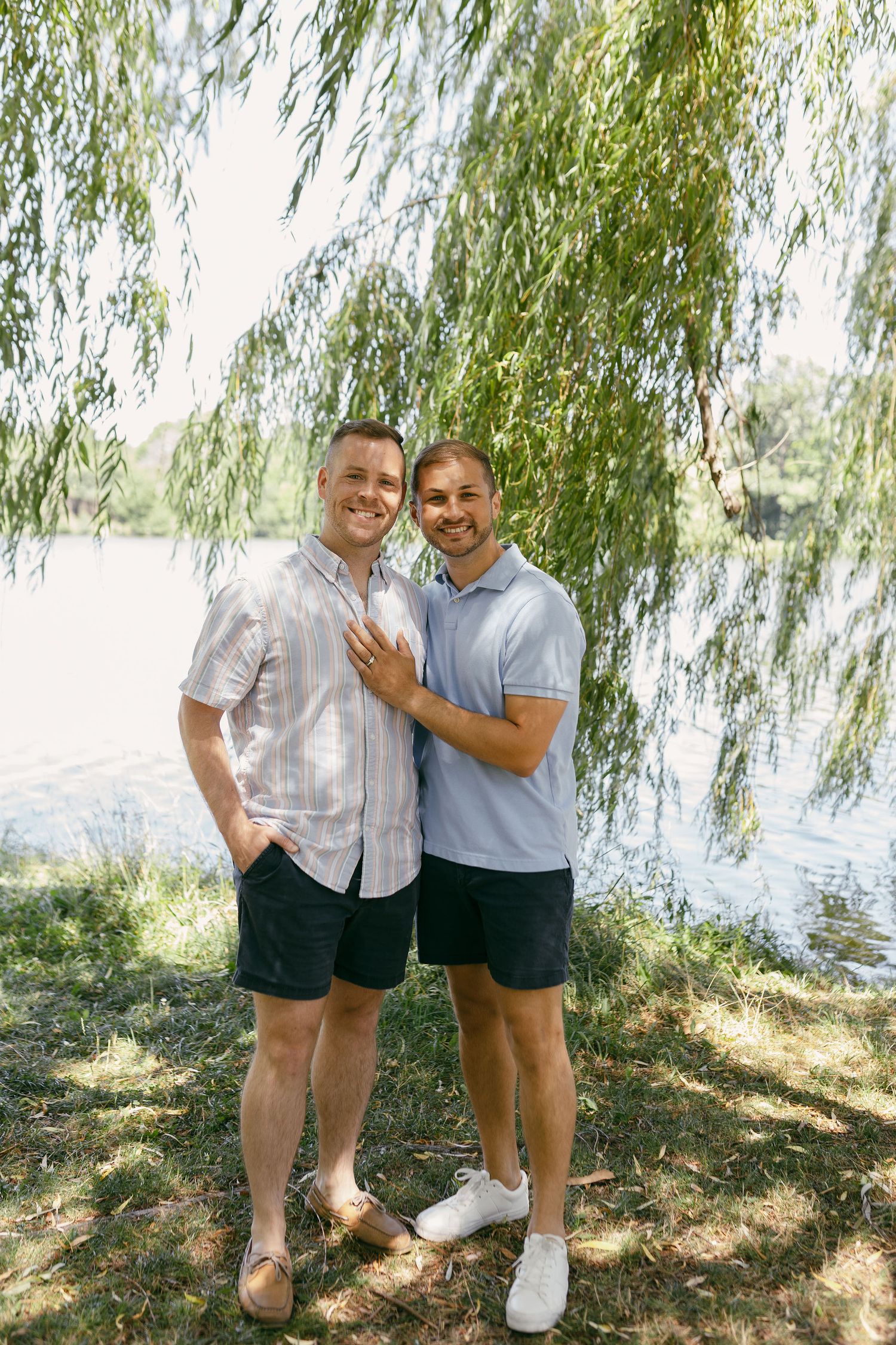 A happy couple poses under a weeping willow tree near a lake.