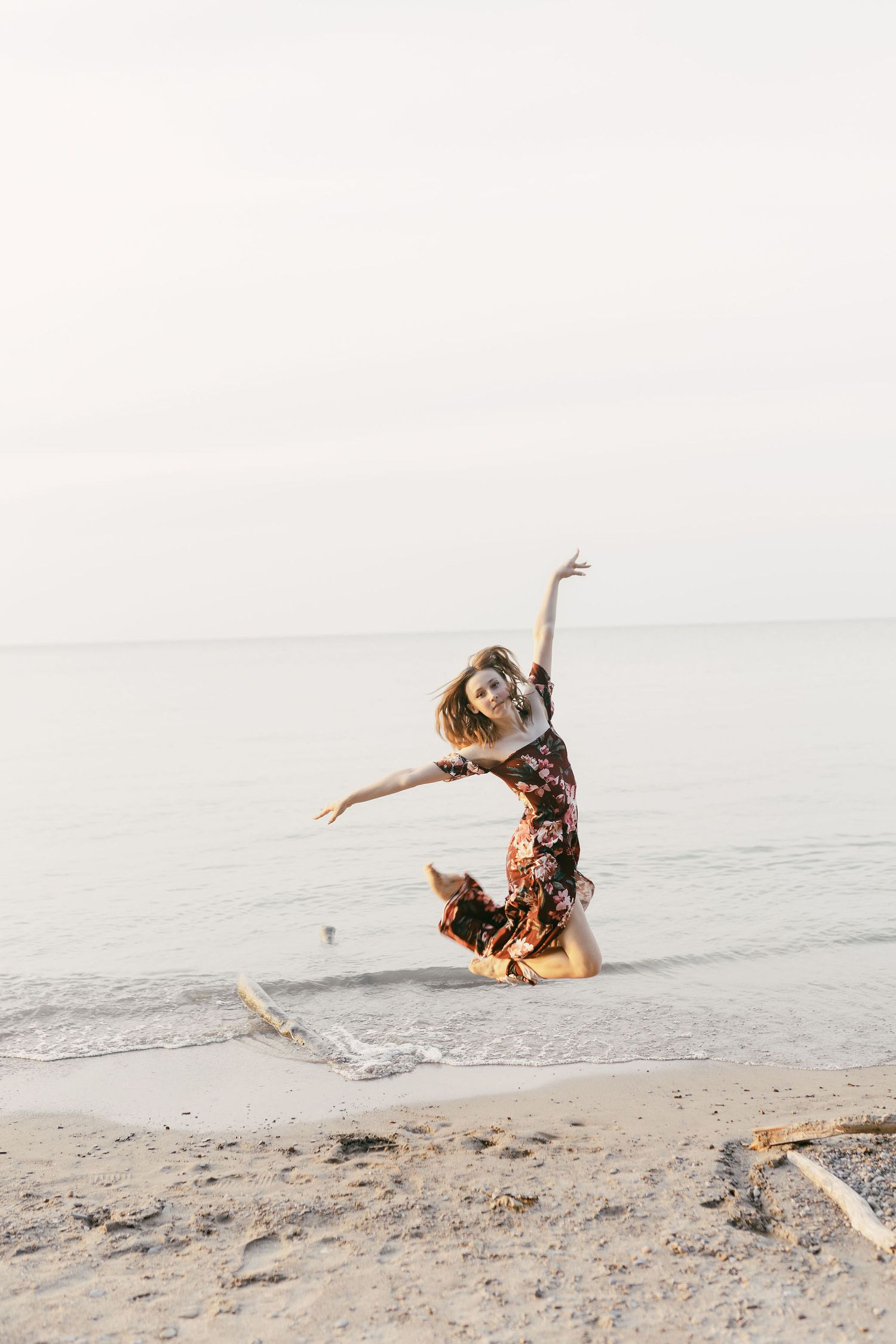 Woman in floral dress joyfully jumping on a sandy beach with calm ocean waters in the background.