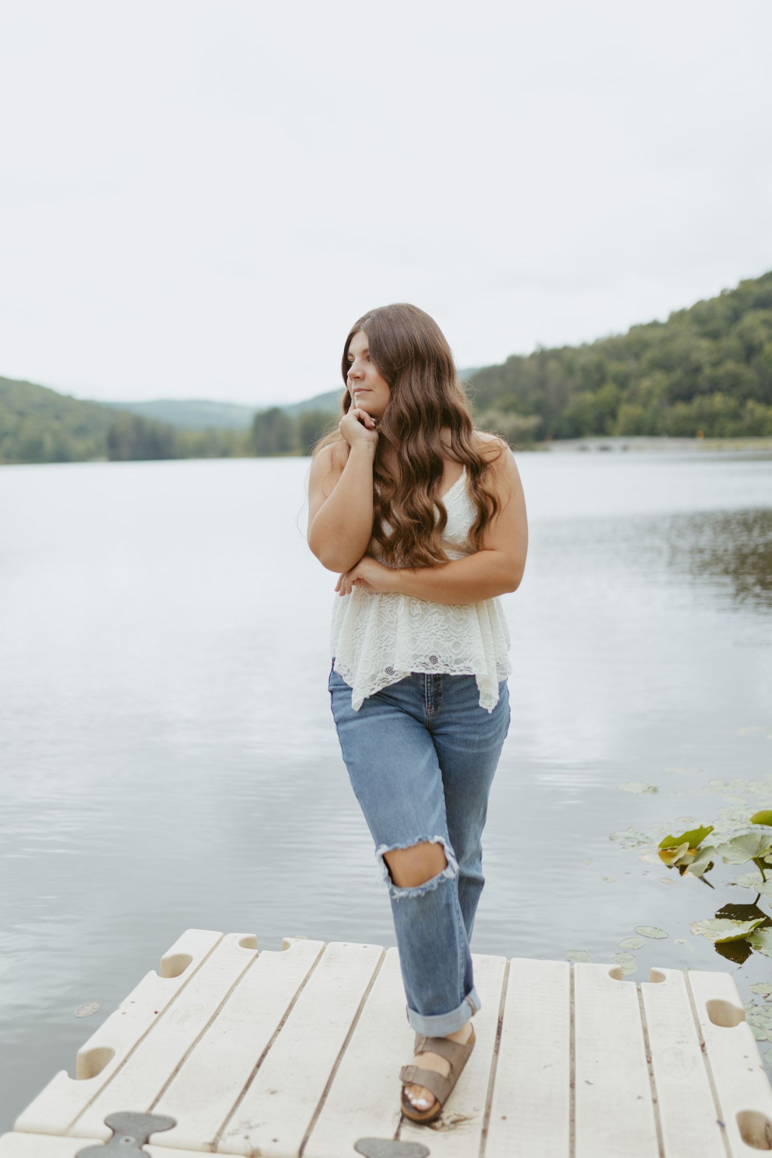 Young woman in white lace top and ripped jeans poses on a wooden dock overlooking a serene lake.