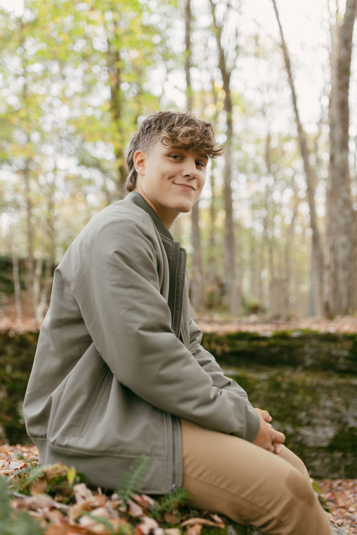 A smiling teenage boy sits on a moss-covered rock in an autumn forest, wearing a green bomber jacket.