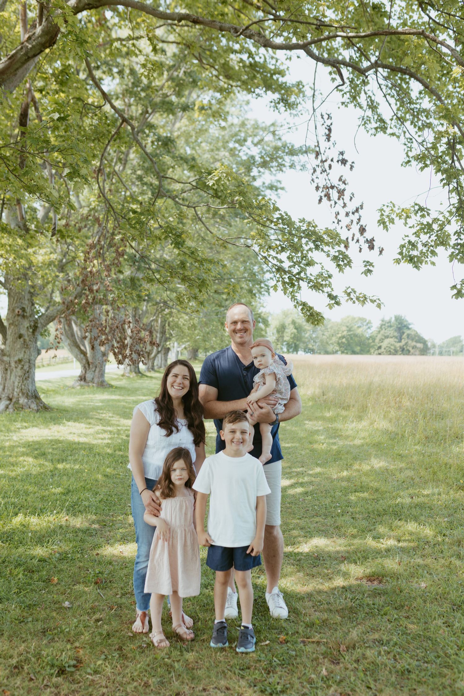 Family of five posing outdoors under large trees in a green park, parents with three young children smiling.