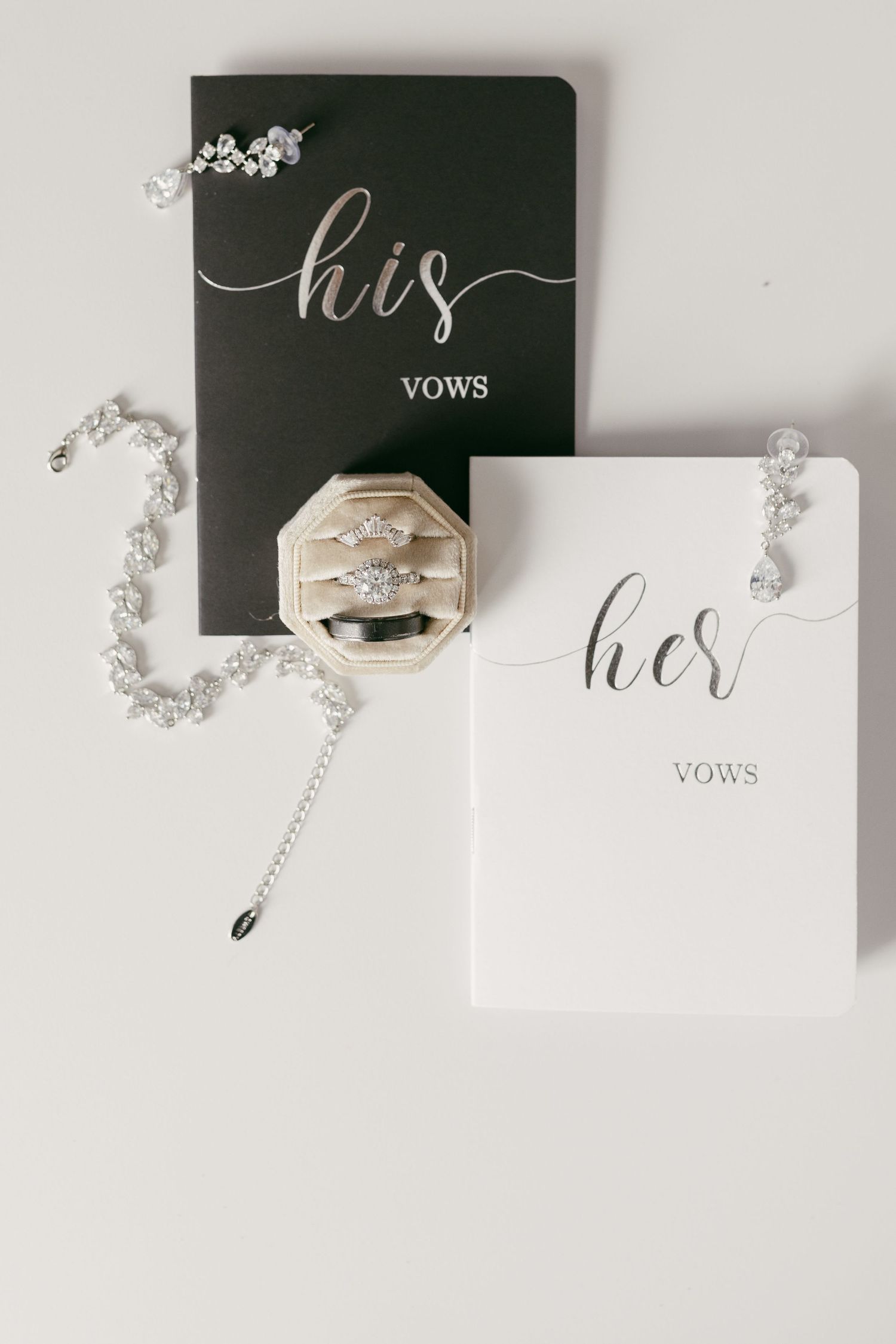 Wedding flat lay featuring his and her vow books, diamond rings in velvet box, and crystal jewelry.
