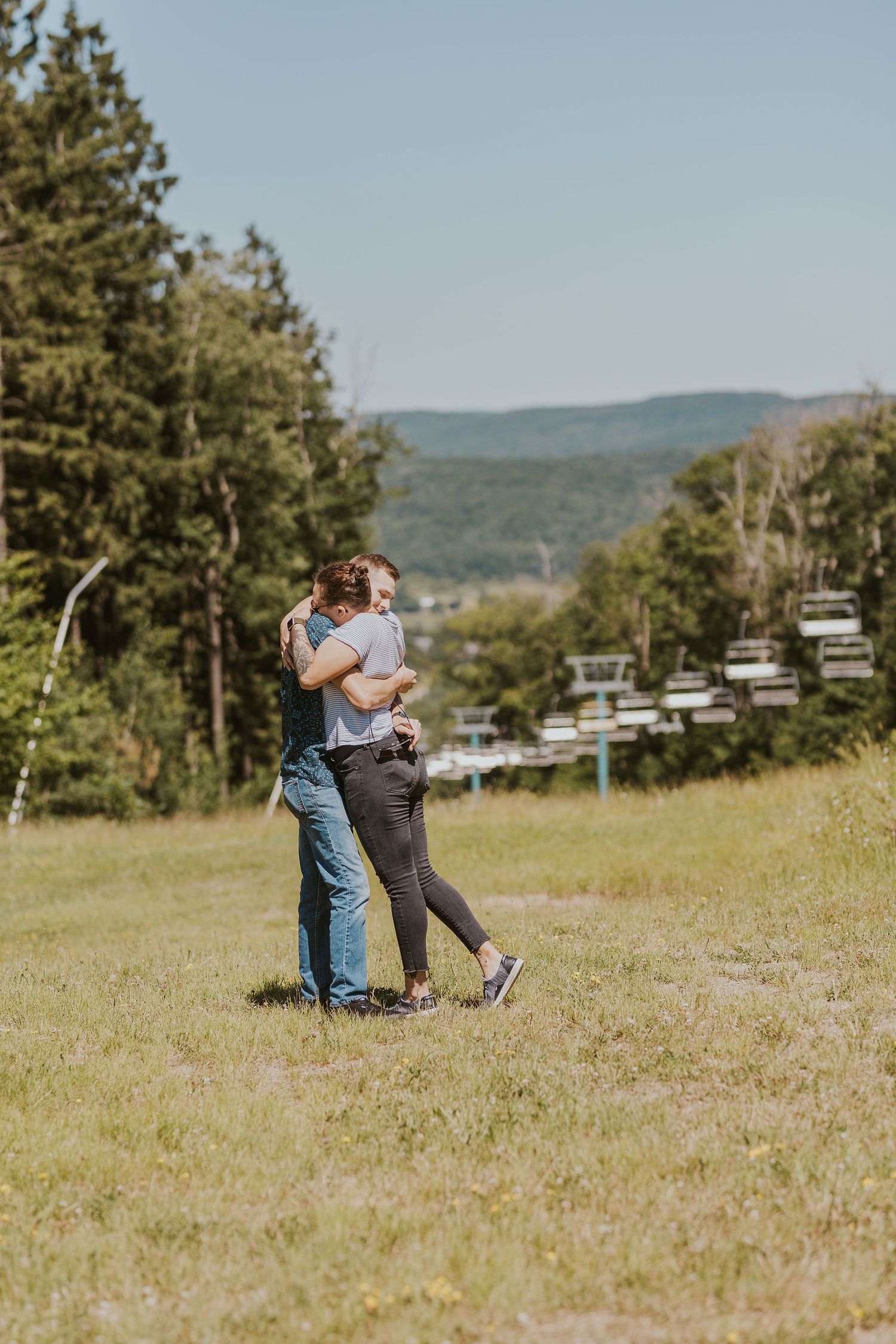 Couple embracing on a grassy hillside near ski lifts, surrounded by lush green trees and mountains on a sunny day.