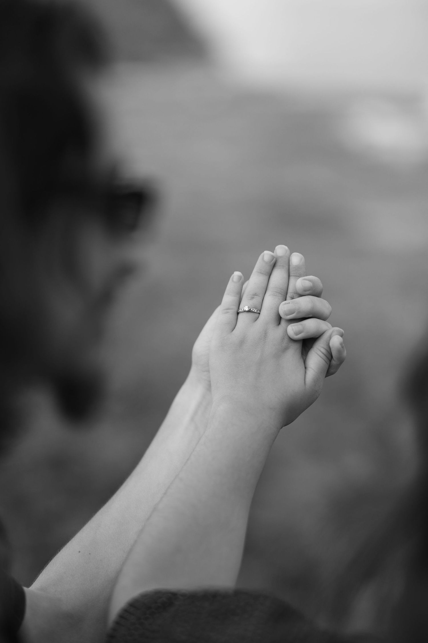 Black and white photo of two hands held together, one wearing a delicate ring, against blurred background.
