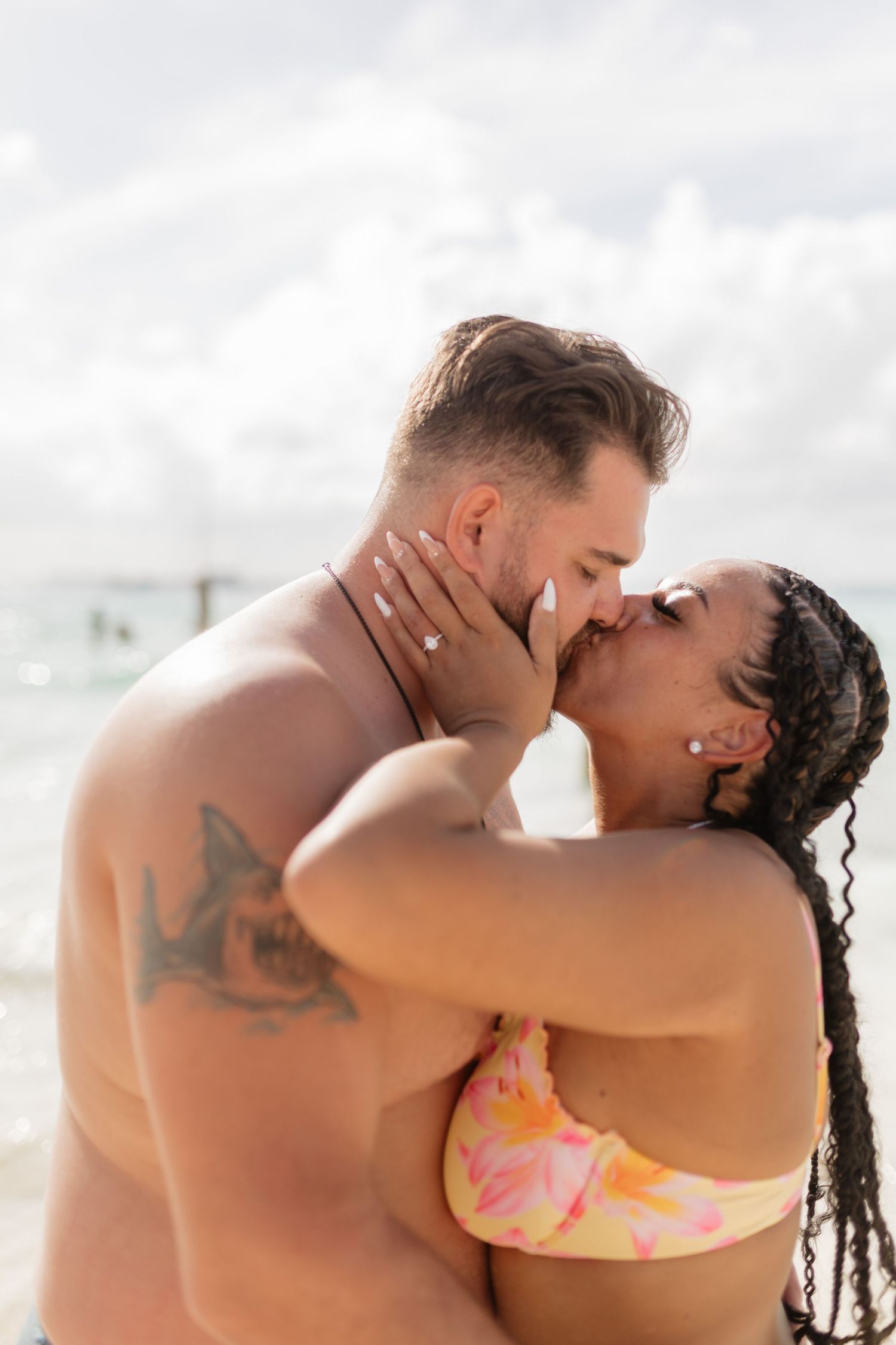 Couple sharing a romantic kiss on the beach, woman in floral bikini with braids, man with tattoo, ocean backdrop.