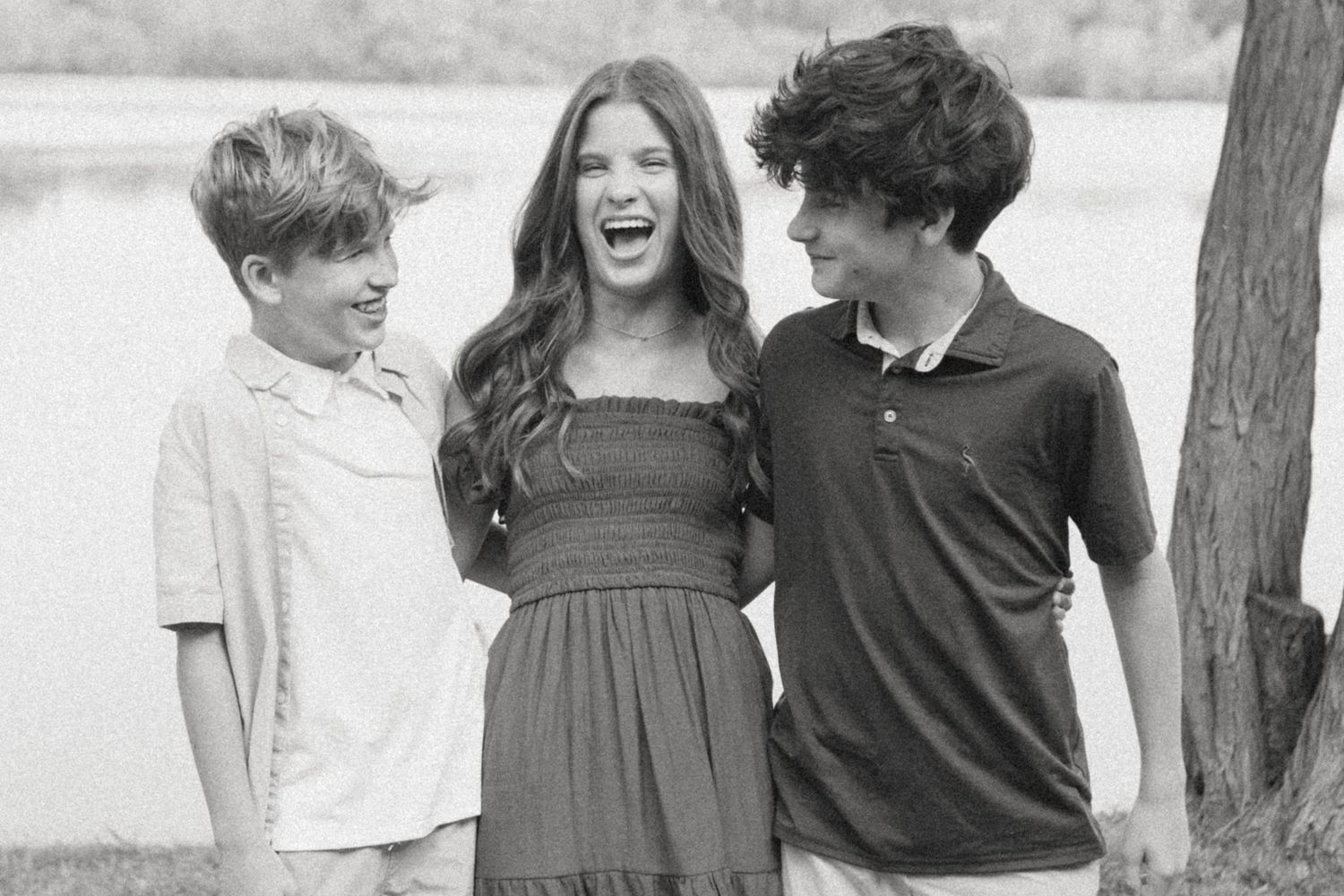 Three smiling teenagers posing together outdoors near a tree and lake in a black and white photo.