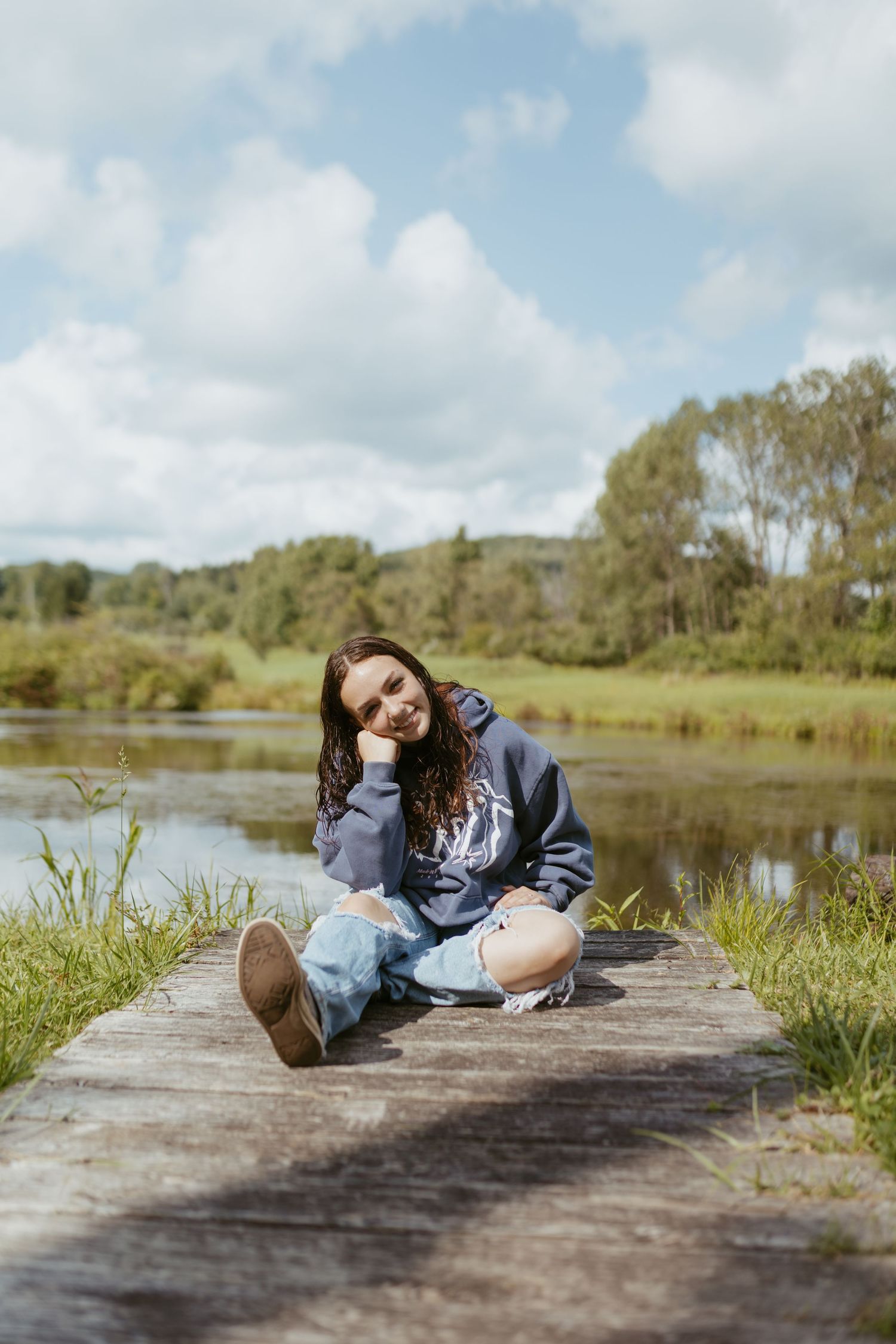 Young woman in blue hoodie and ripped jeans sitting on wooden dock by a serene river, smiling outdoors.
