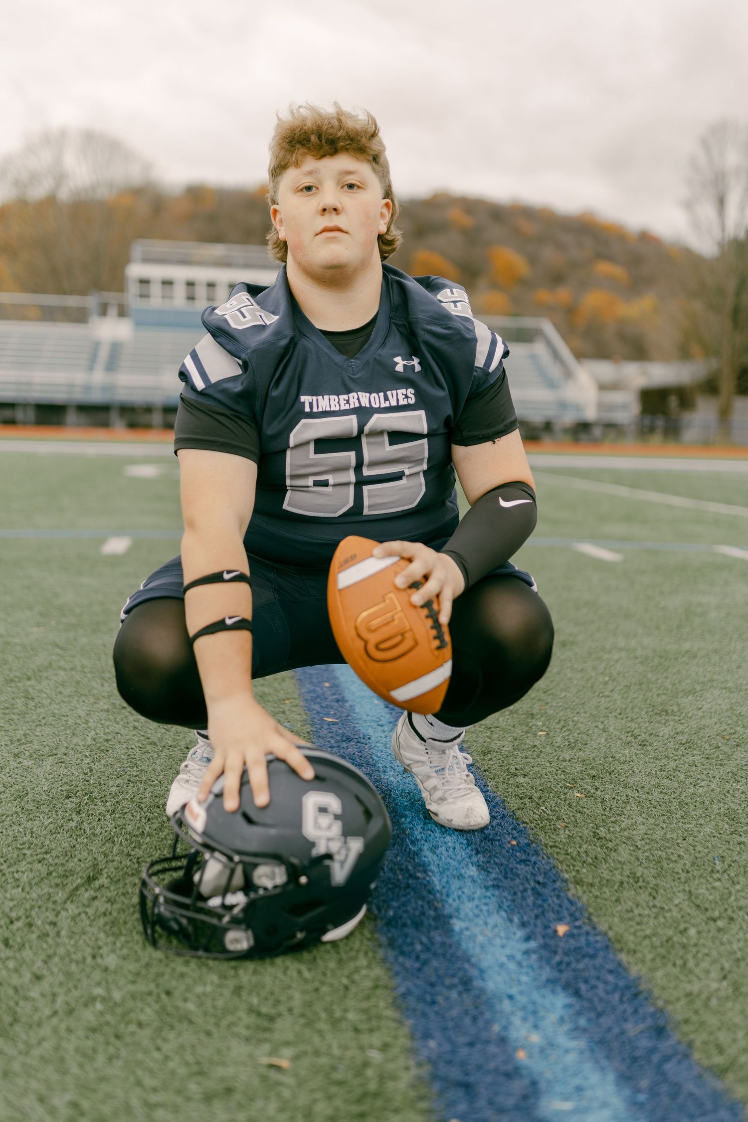 Young football player in Timberwolves #65 jersey crouching on field holding ball with helmet beside him.