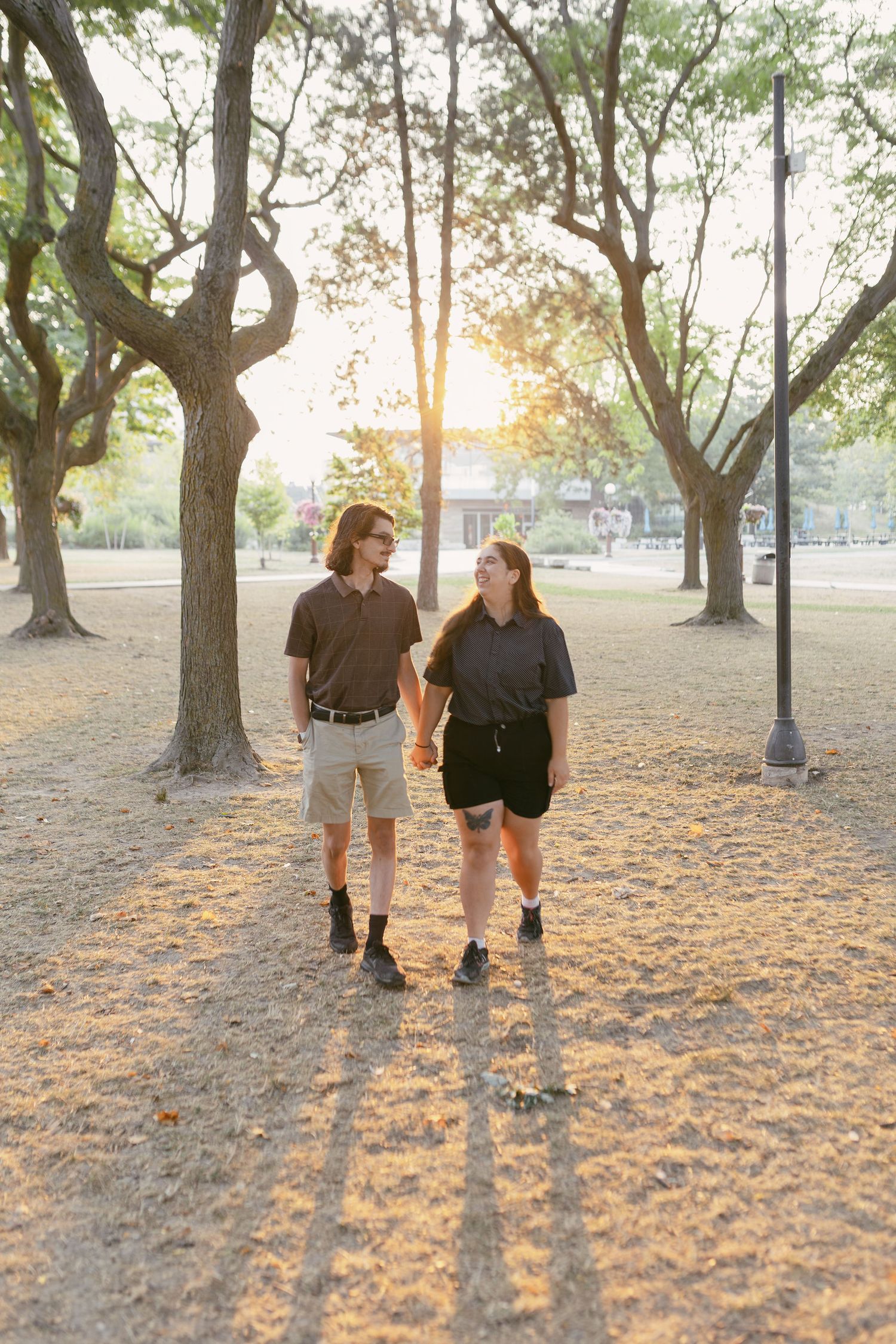 A couple holds hands and walks through a sunlit park with tall trees casting long shadows on the ground.