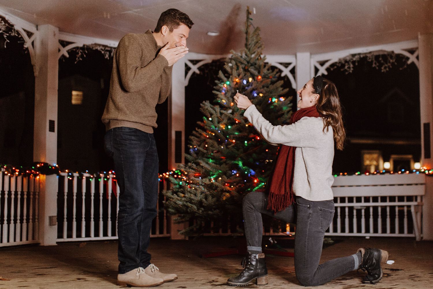 Woman proposes to man on one knee outdoors near decorated Christmas tree with holiday lights at night.