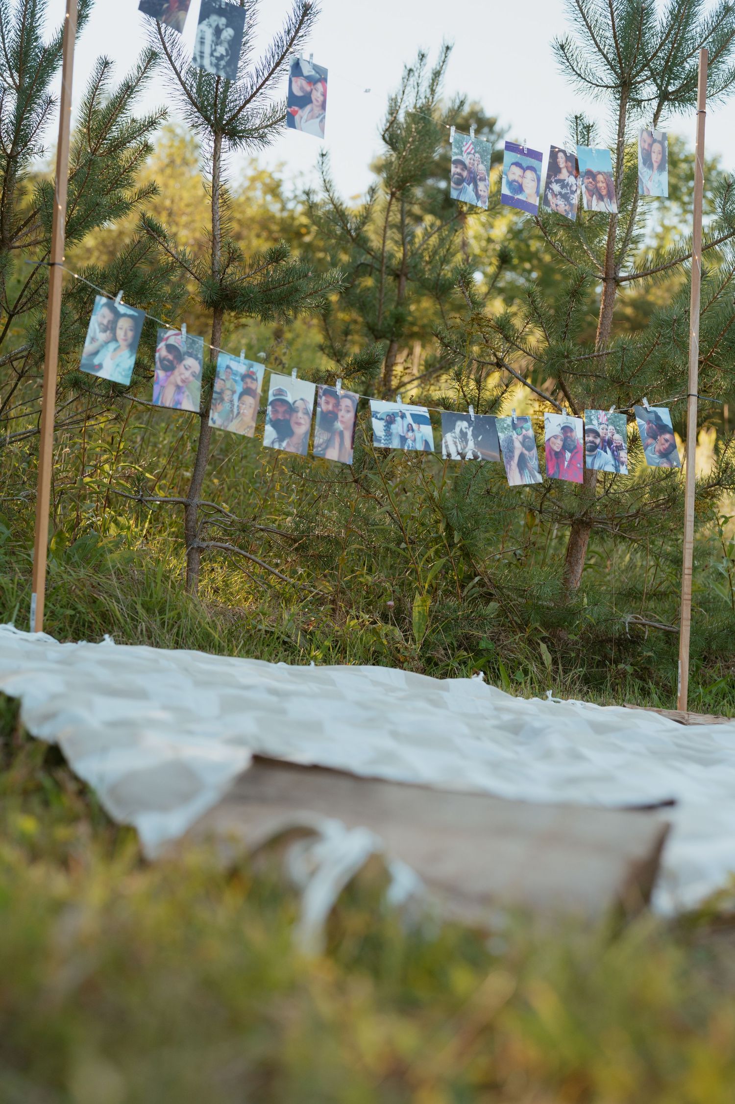 Photos strung on a garland between wooden poles in a forest clearing with a white blanket on the ground.