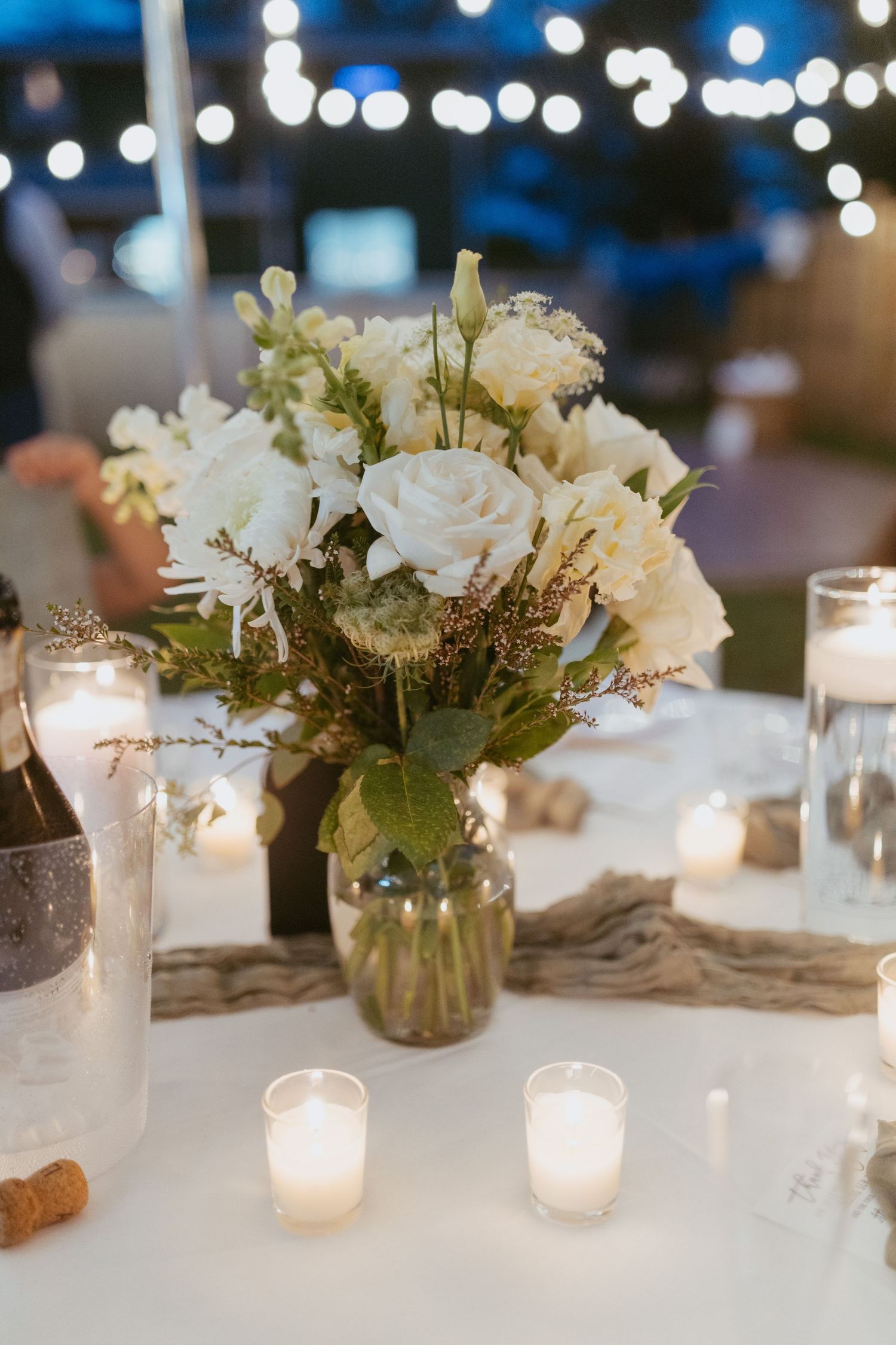 Elegant wedding reception centerpiece with white roses, candles, and greenery in a glass vase on a white tablecloth.