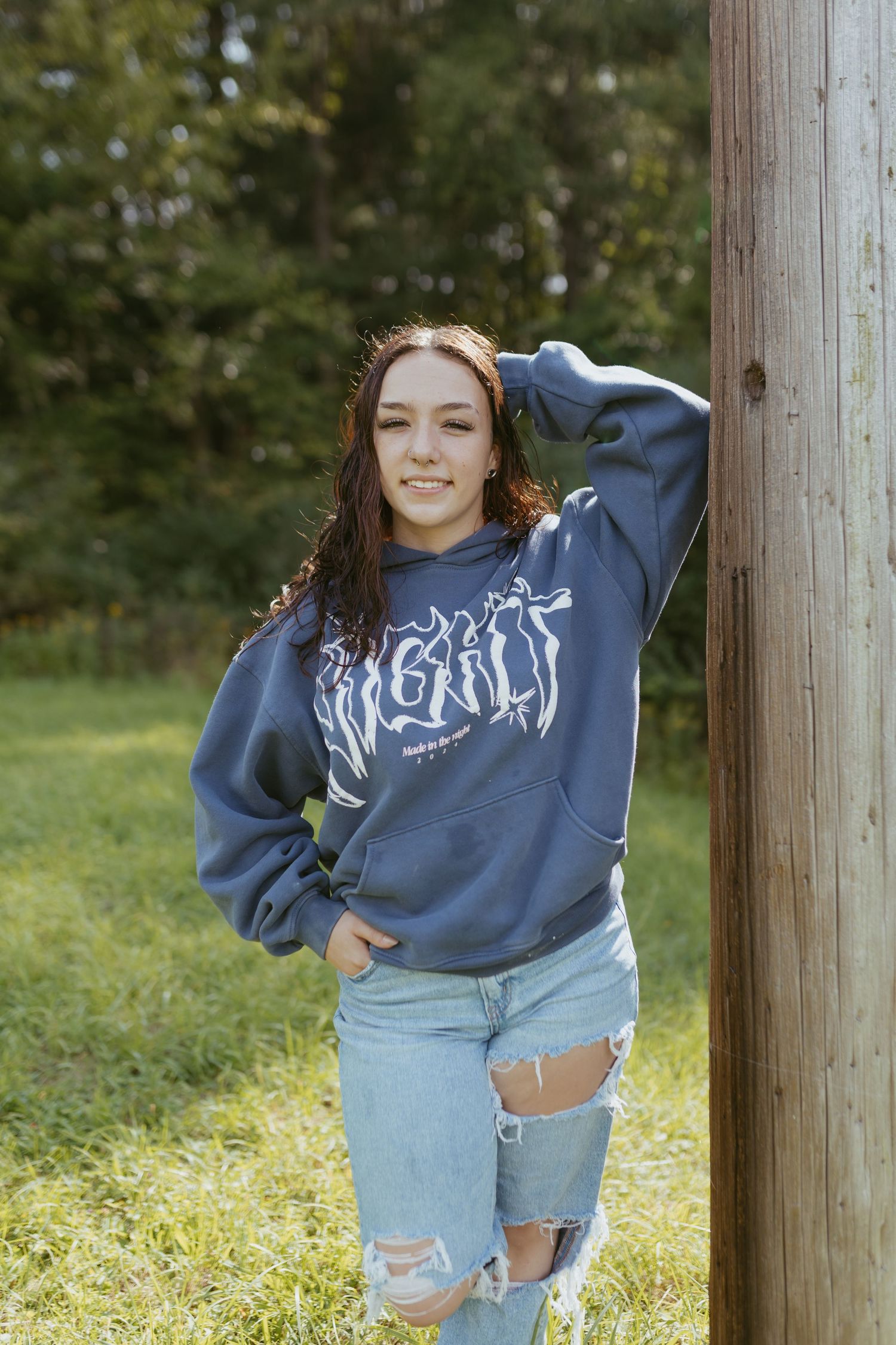 Young woman in navy graphic hoodie and ripped jeans leaning against wooden post outdoors with green trees behind her.