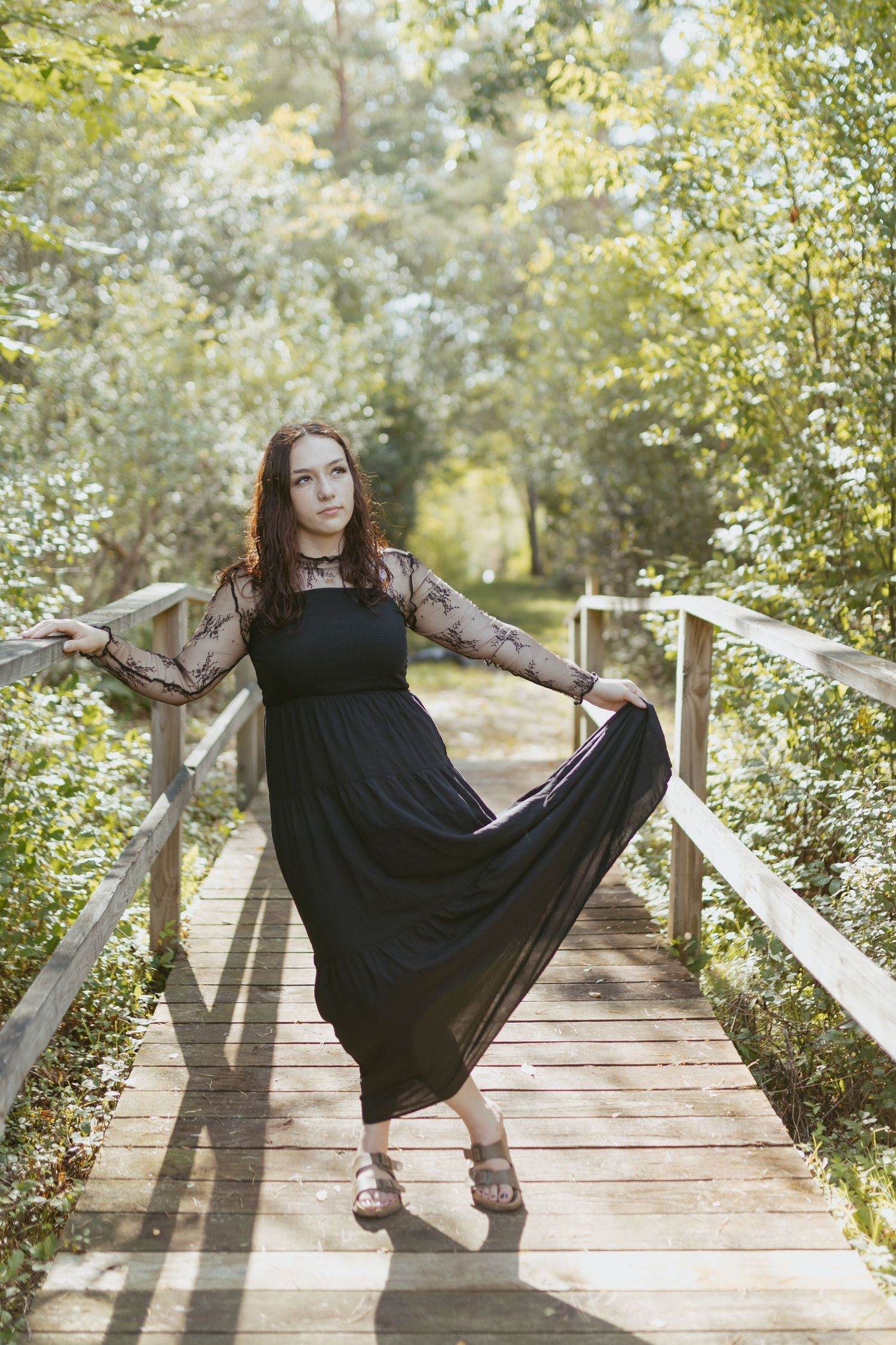 Woman in black lace maxi dress posing on wooden bridge surrounded by lush green trees in sunlight.