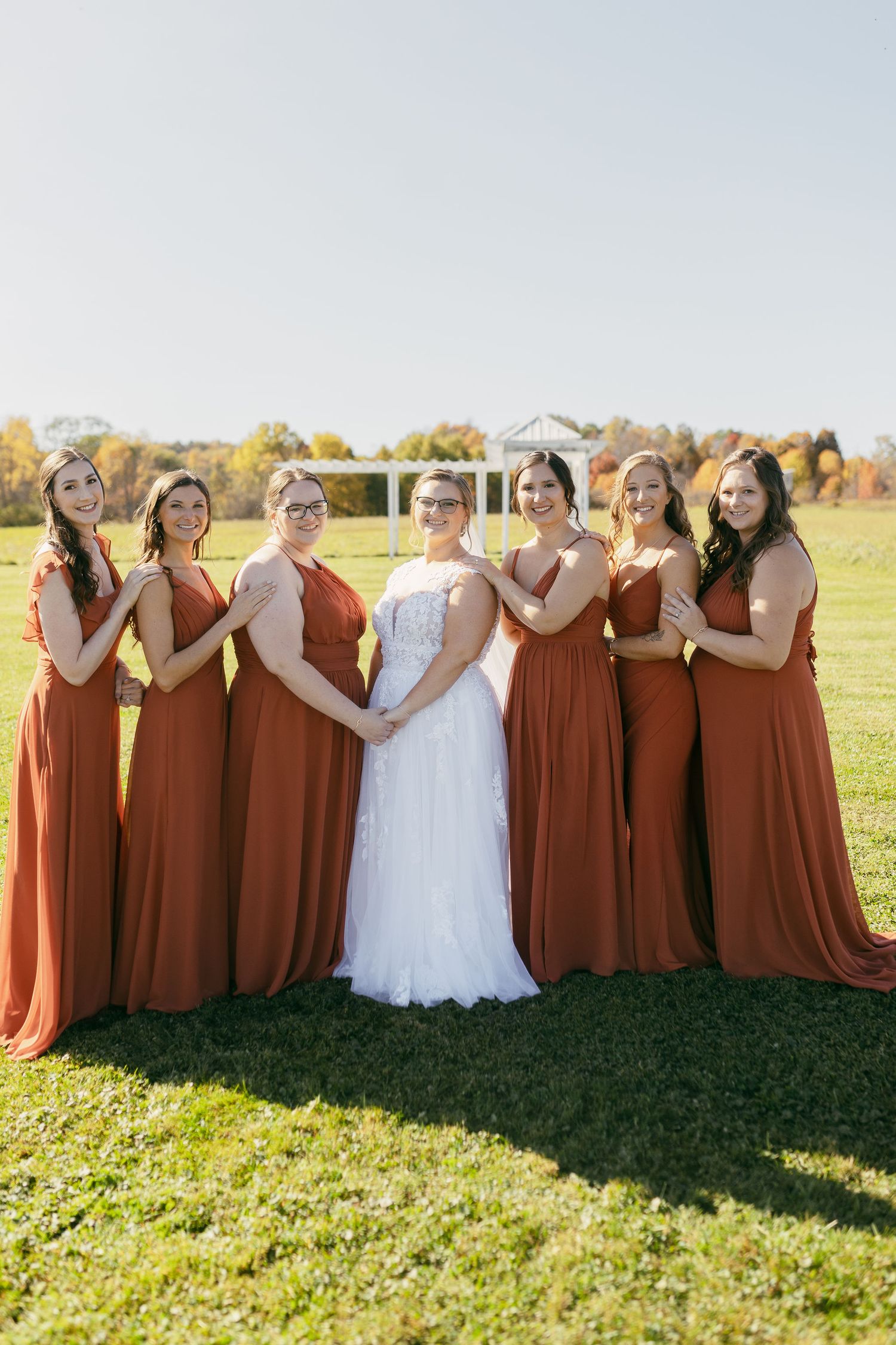 Bridal party in rust-colored dresses standing together on grass during fall wedding.