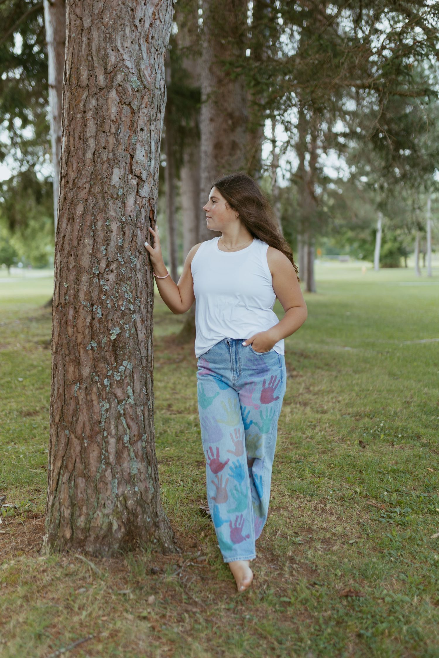 Woman in white tank top and colorful hand-print jeans leaning against a tree in a park.