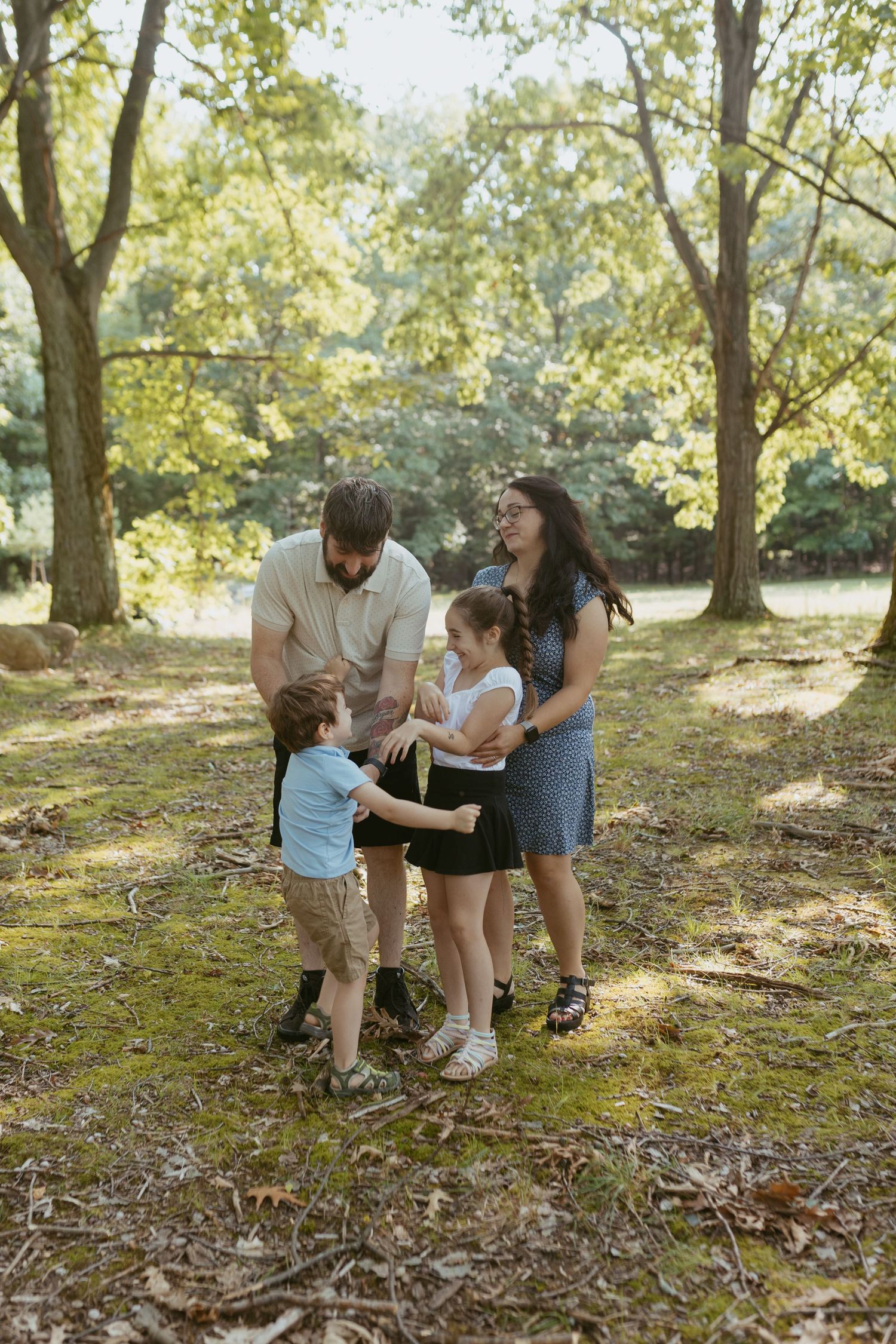 Family of four enjoying a sunny day in a lush green forest park, with parents watching their two young children play.