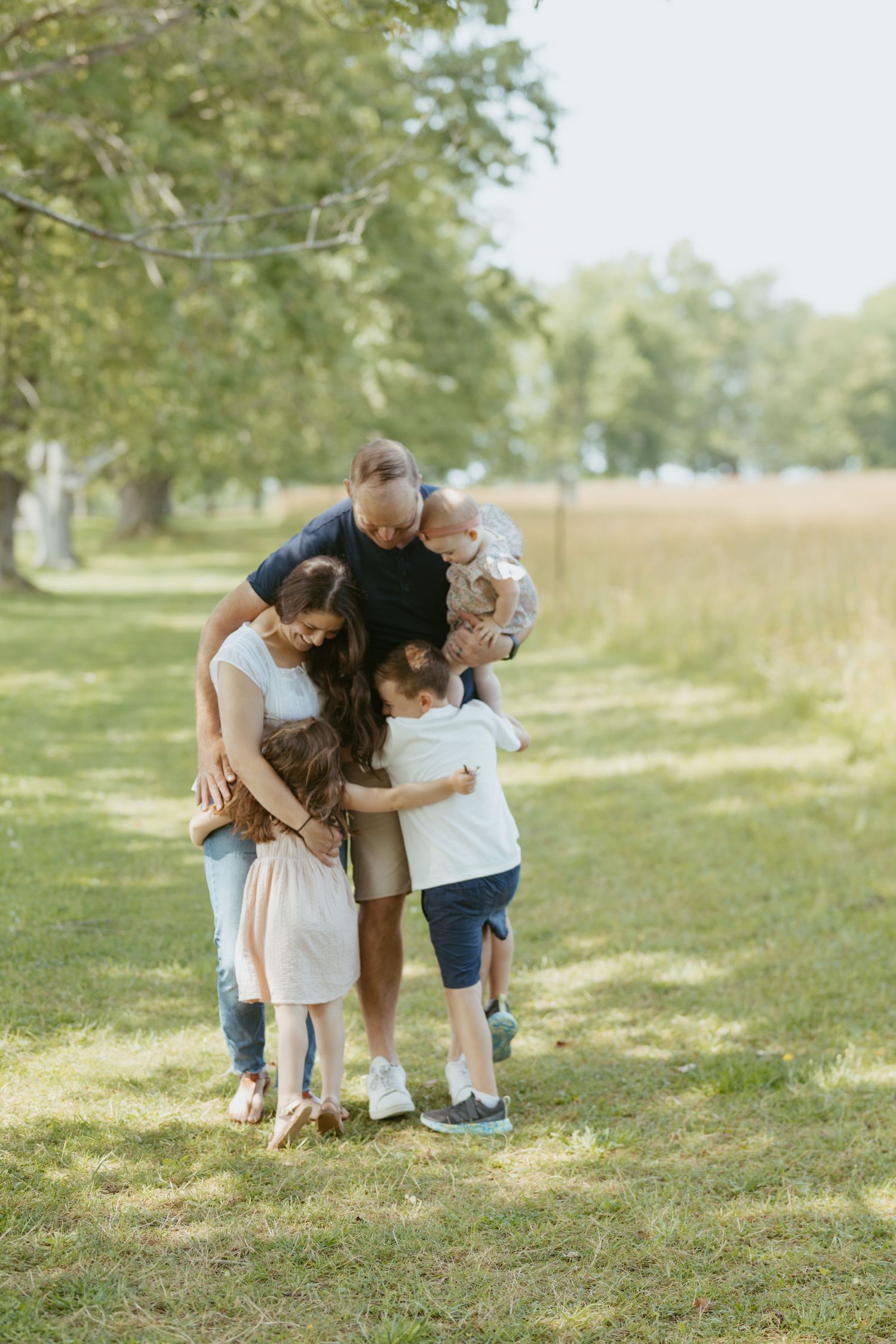 A loving family shares a group hug outdoors in a sunny green park, with parents and three young children embracing.