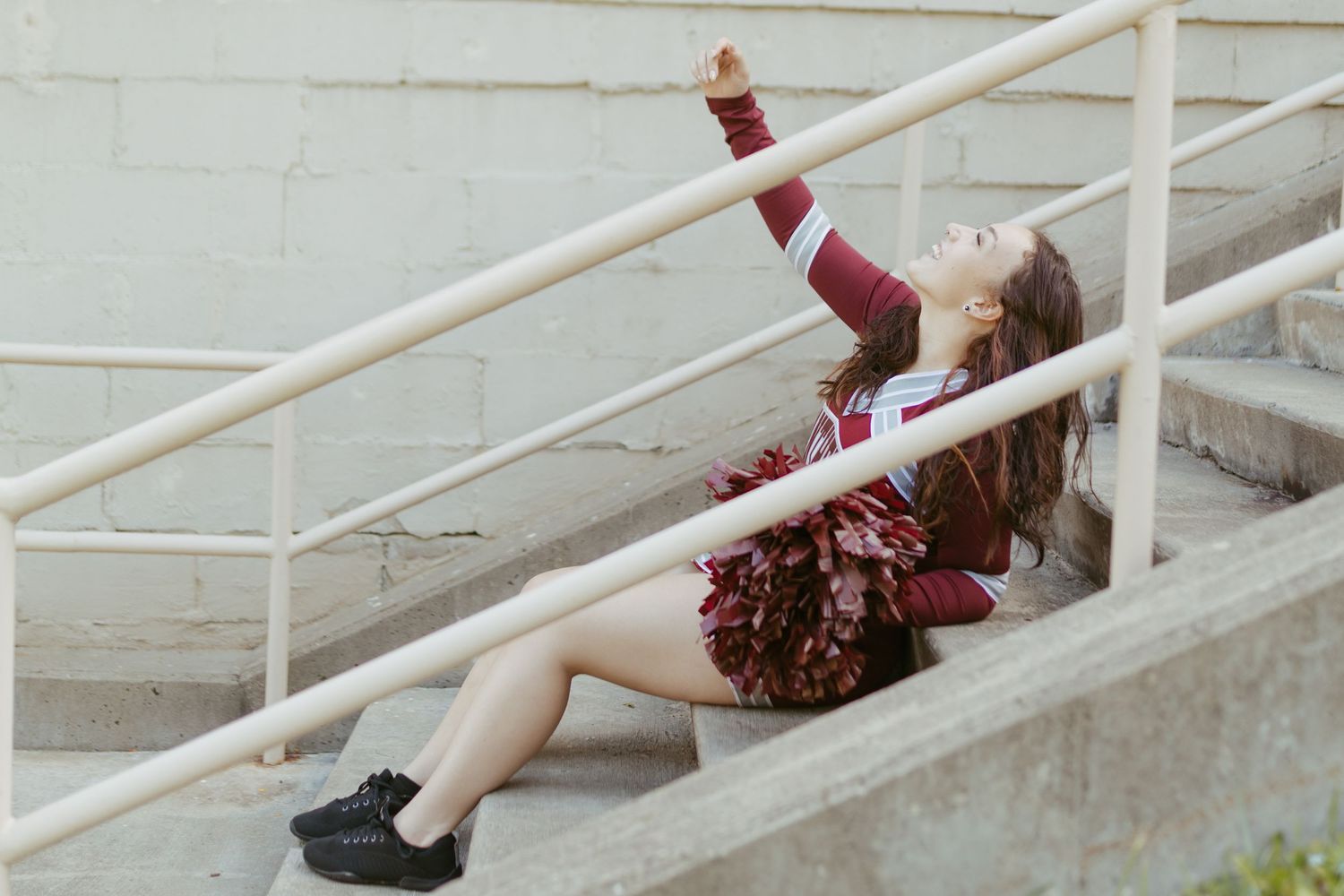 Cheerleader in maroon uniform sitting on concrete steps holding pom-poms with one arm raised.