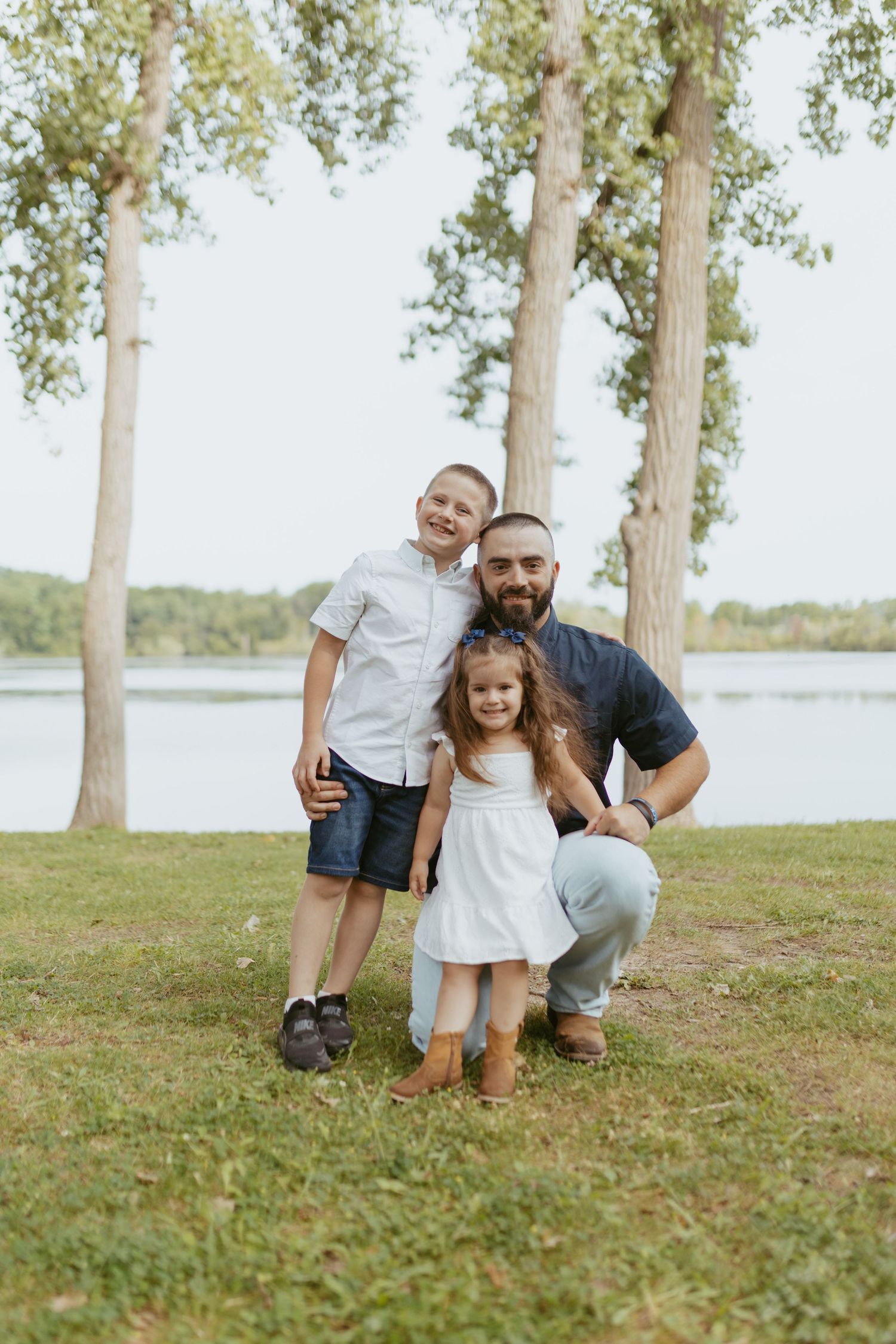 A father crouches with his two young children near a lakeside park, surrounded by tall trees on a warm day.