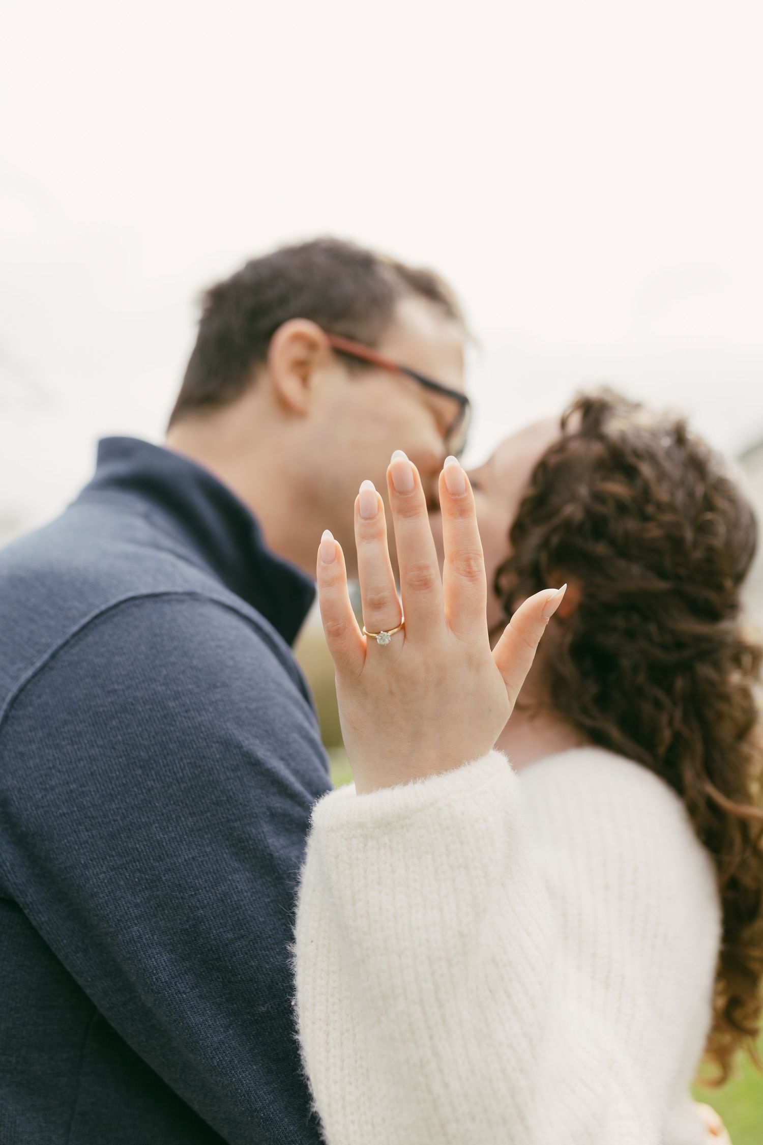 Couple kissing while woman shows off engagement ring on outstretched hand, wearing white sweater outdoors.
