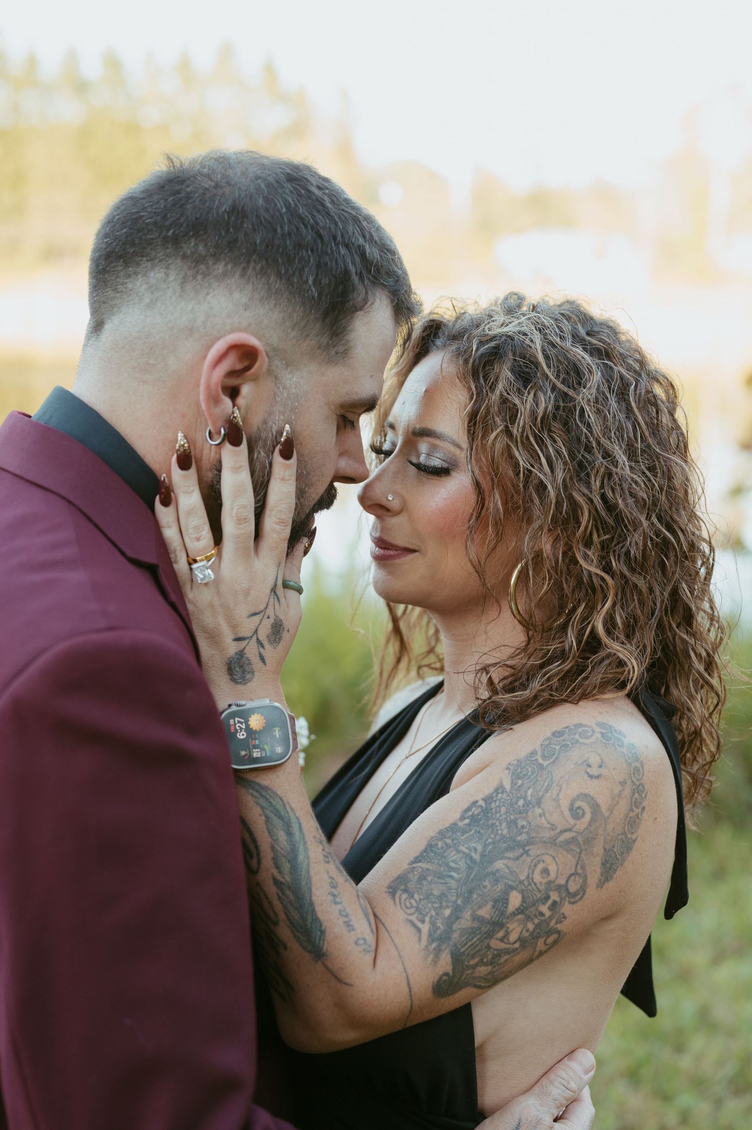 Tattooed couple sharing an intimate moment, woman in black dress touching man's face, golden hour outdoor engagement photo.