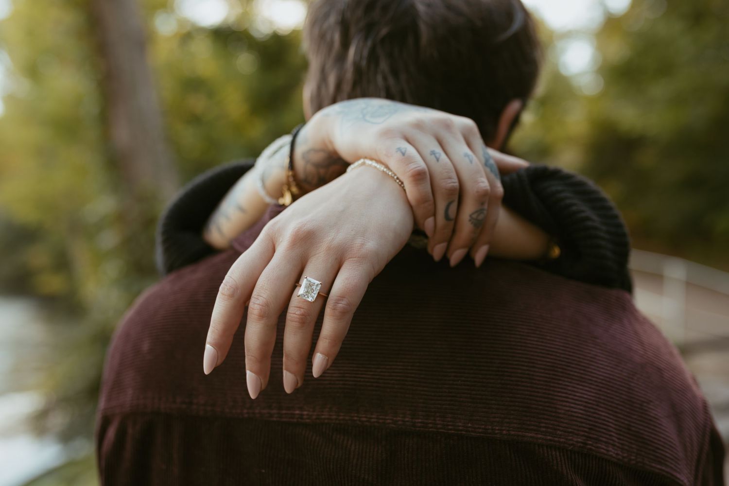 Couple embracing outdoors, woman's hands showing engagement ring and gold bracelets resting on man's shoulders.