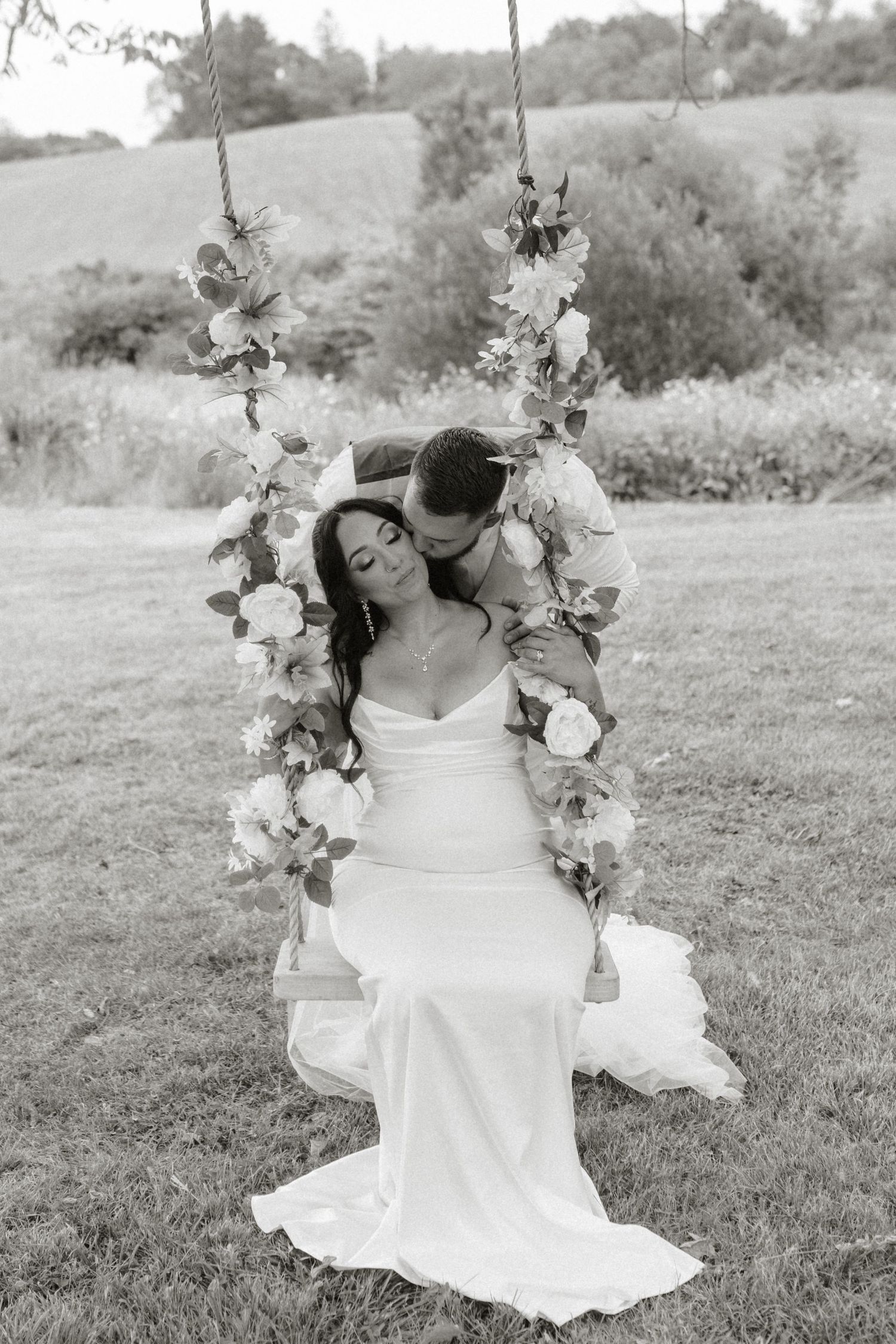 Black and white photo of a bride sitting on a floral swing while the groom kisses her cheek outdoors.