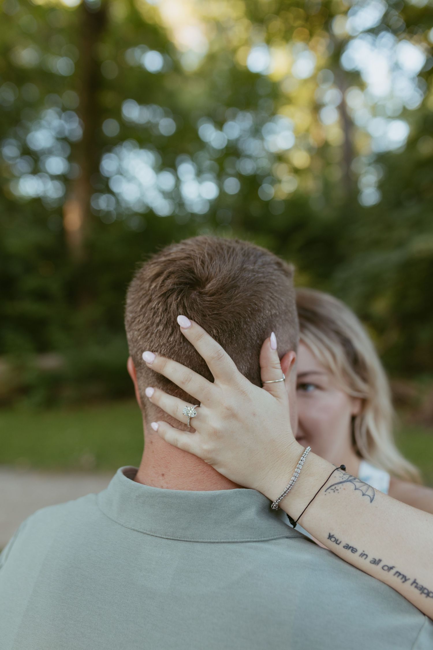 Couple embracing outdoors, woman's hand with engagement ring resting on man's neck, tattoo visible on her arm.