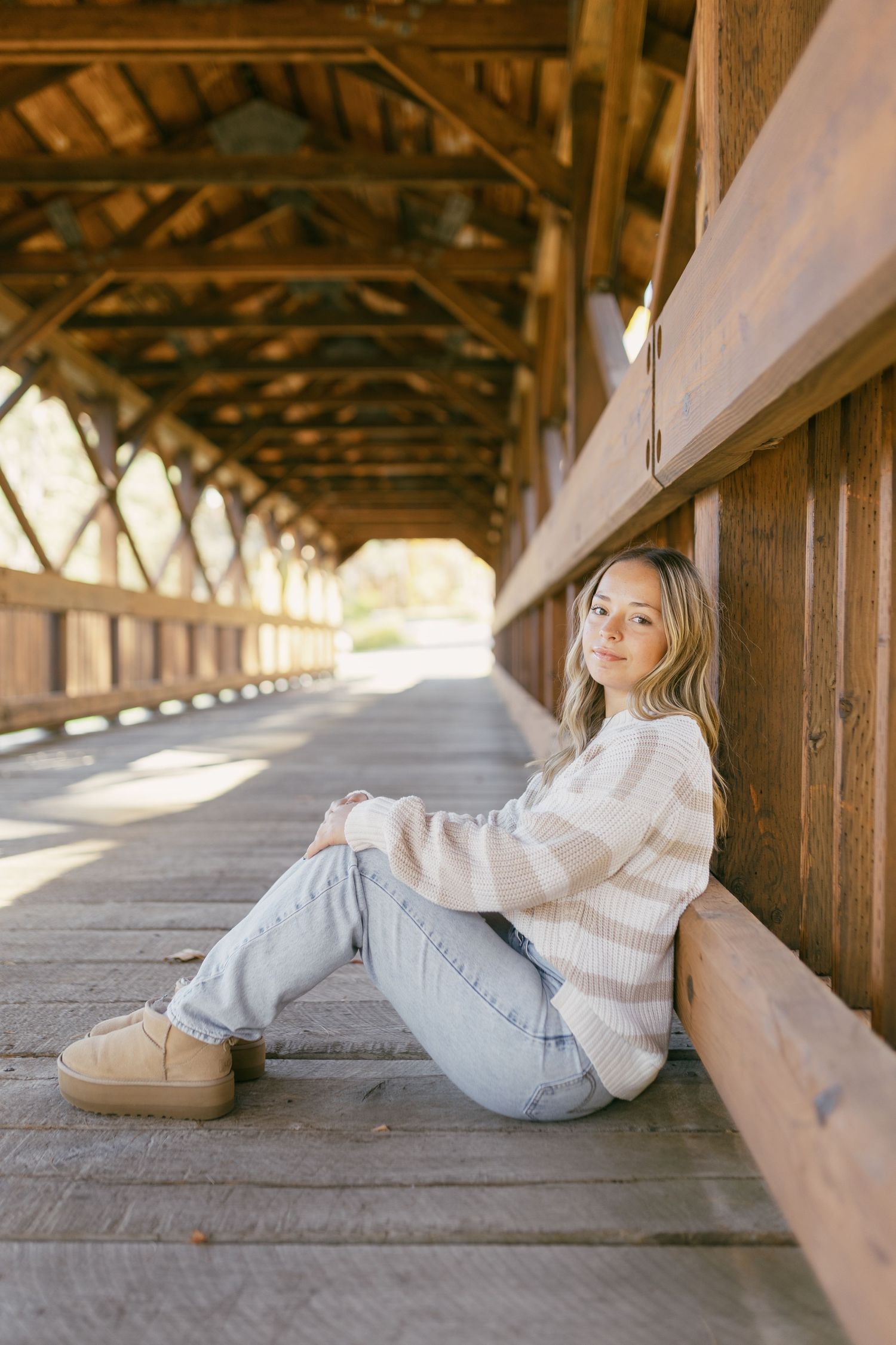 Young woman in striped sweater and jeans sitting against wooden wall inside a rustic covered bridge.
