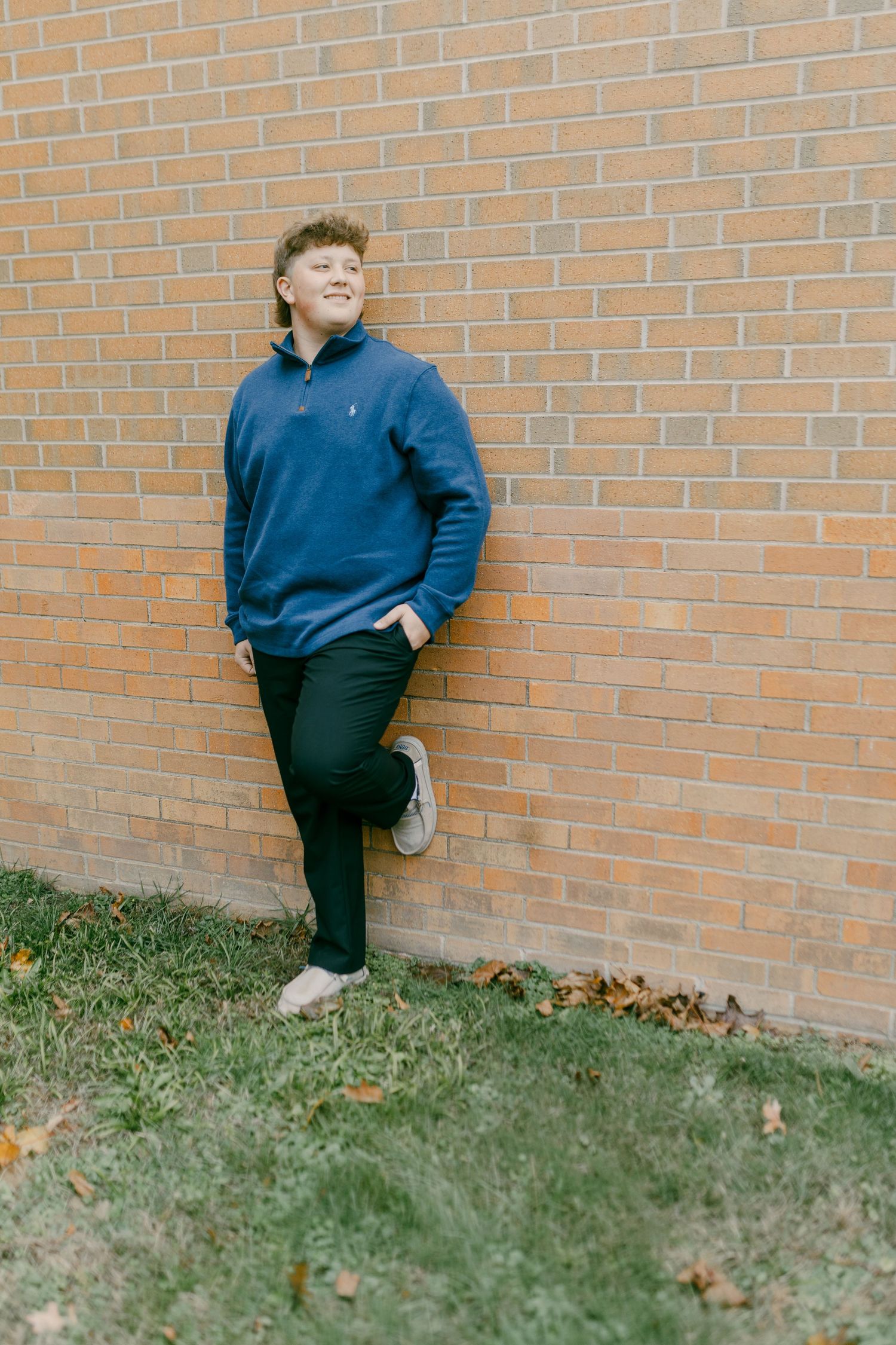 A young man in a blue Polo Ralph Lauren quarter-zip leans against a brick wall outdoors in dark pants and sneakers.