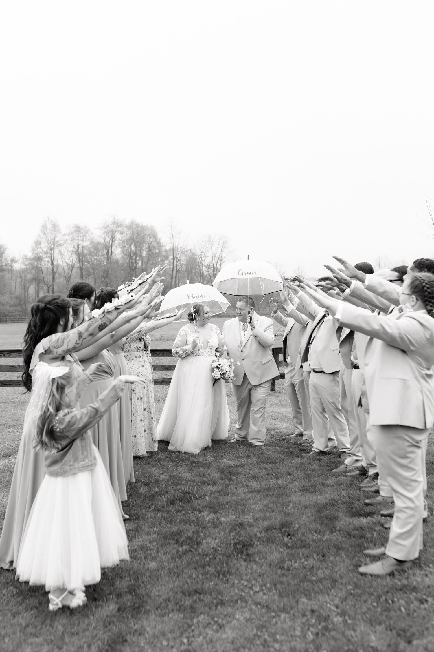 Black and white photo of bride and groom walking through arch of raised arms formed by wedding party outdoors.