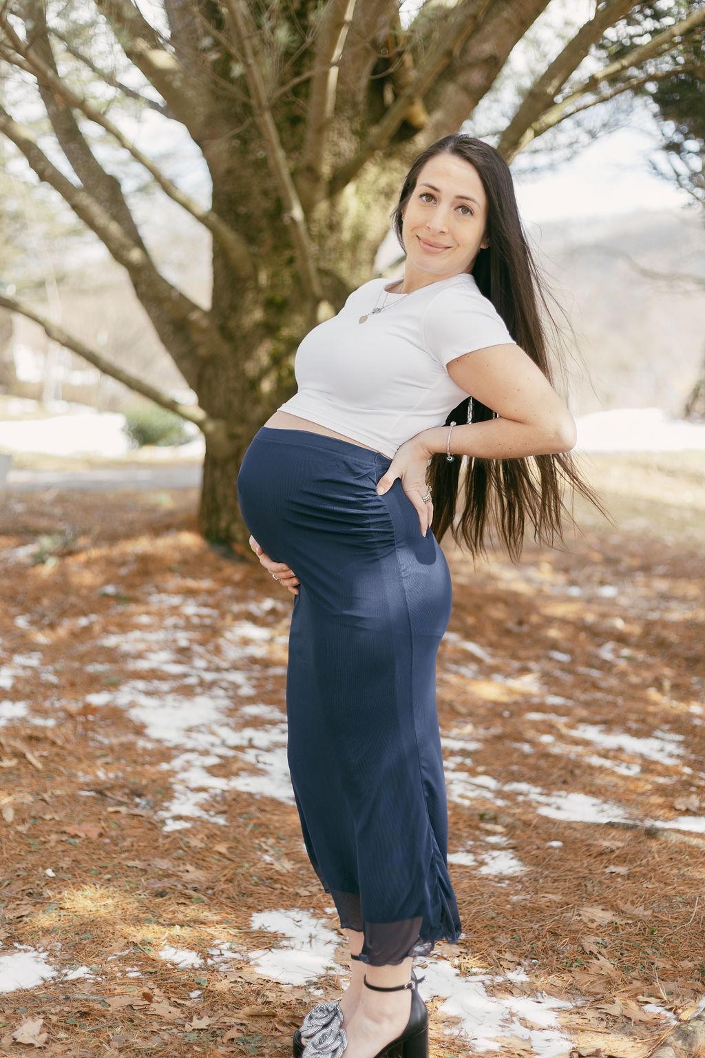 Maternity photos taken under a large tree with patches of snow on the ground.