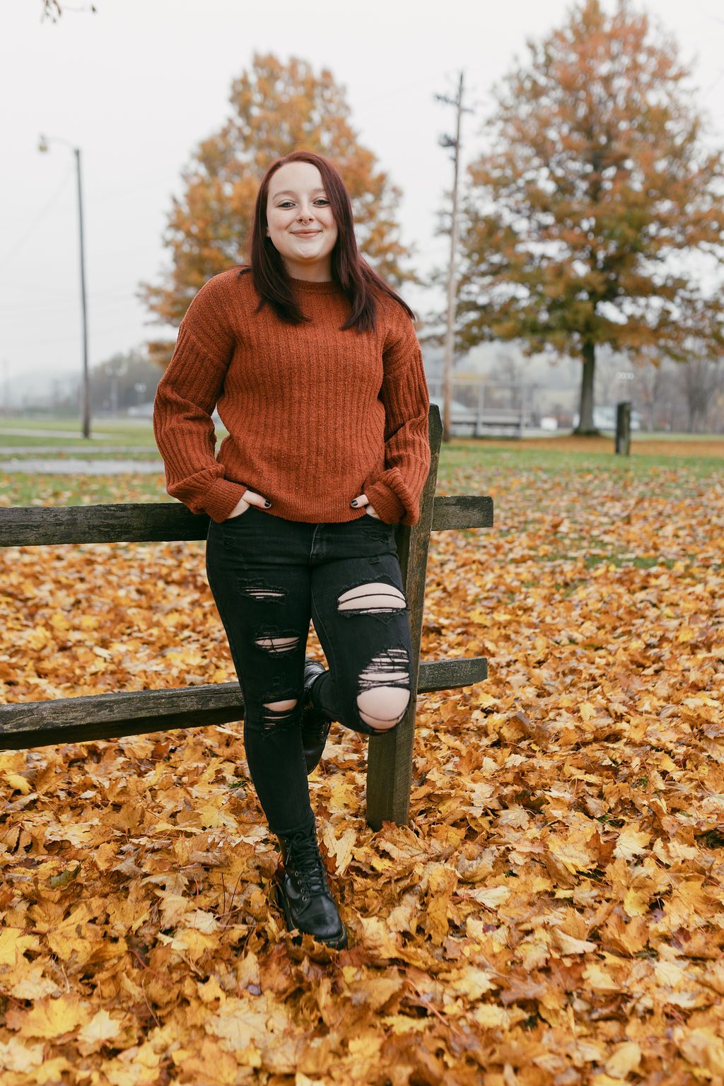 Person in rust-colored sweater sitting on bench surrounded by fallen autumn leaves.