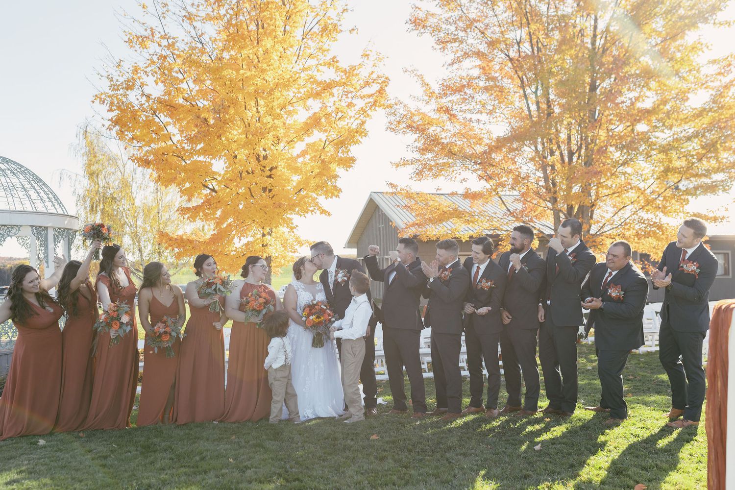 Large wedding party group photo taken outdoors during golden hour with autumn trees in background.