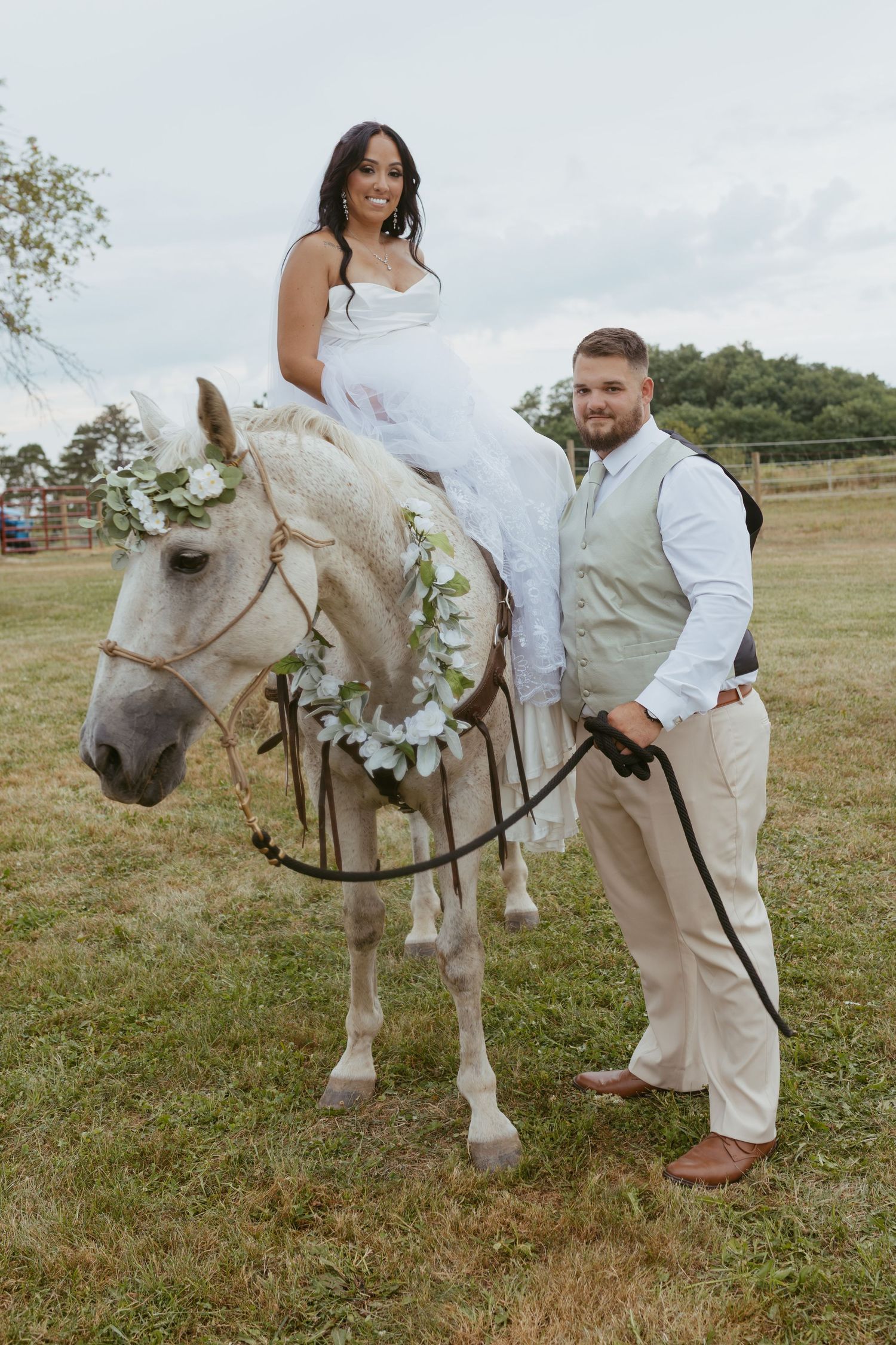 Bride in white gown sits on a decorated white horse held by groom in tan suit at outdoor wedding.