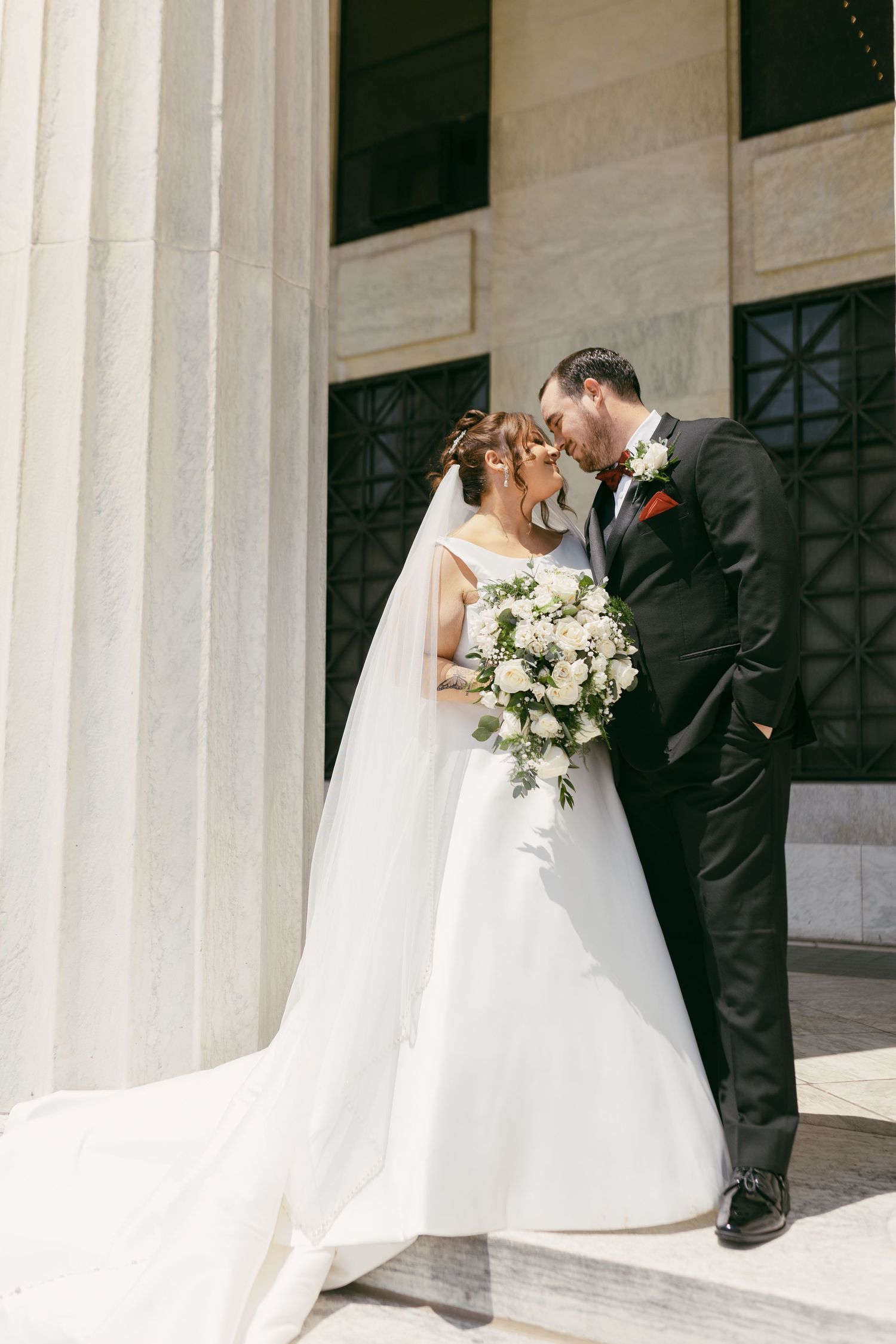 Bride and groom share romantic moment by stone columns, bride holding white floral bouquet.