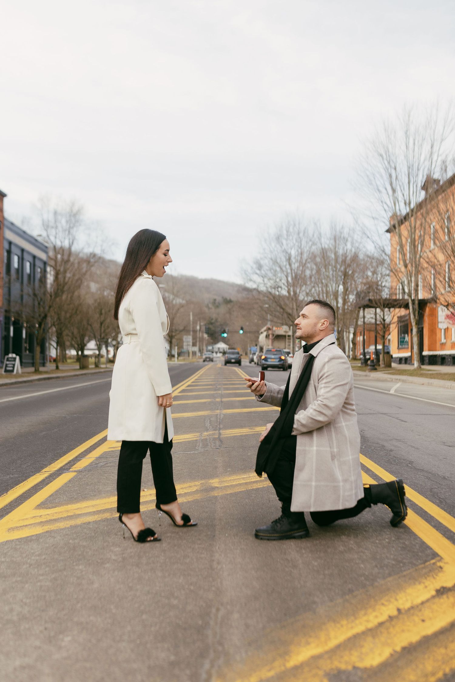 Man proposes on one knee to woman in crosswalk, both wearing elegant coats on a quiet urban street.