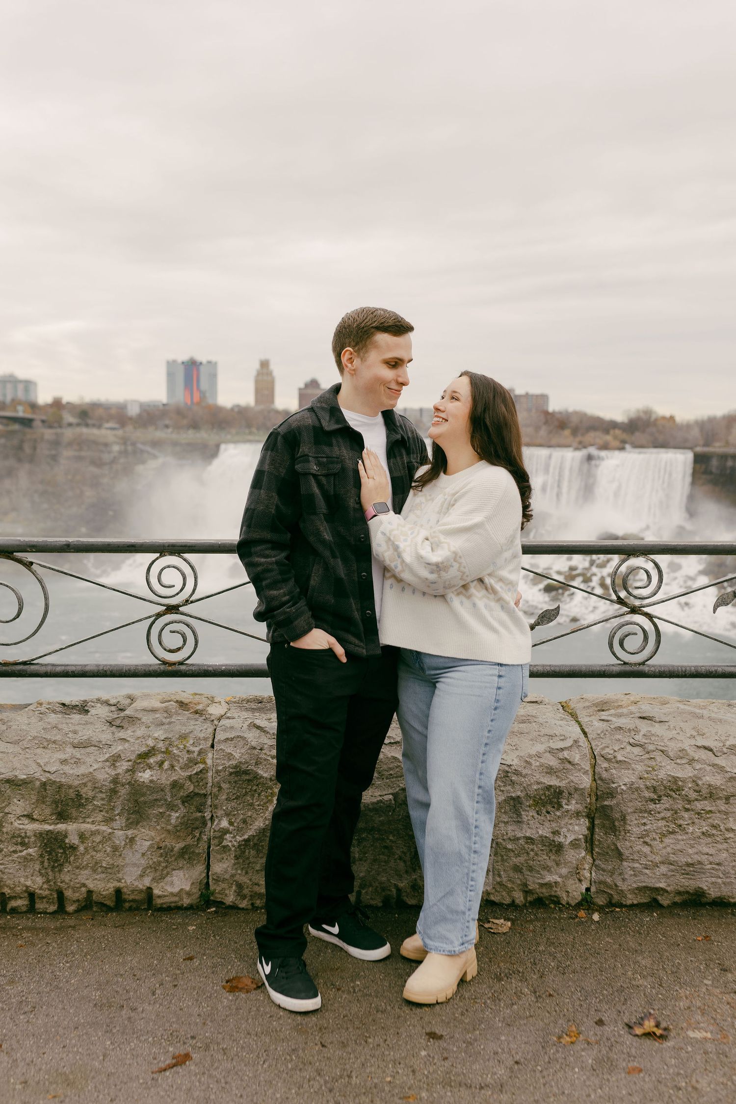 Couple embracing and smiling at each other with Niagara Falls and city skyline in the background.