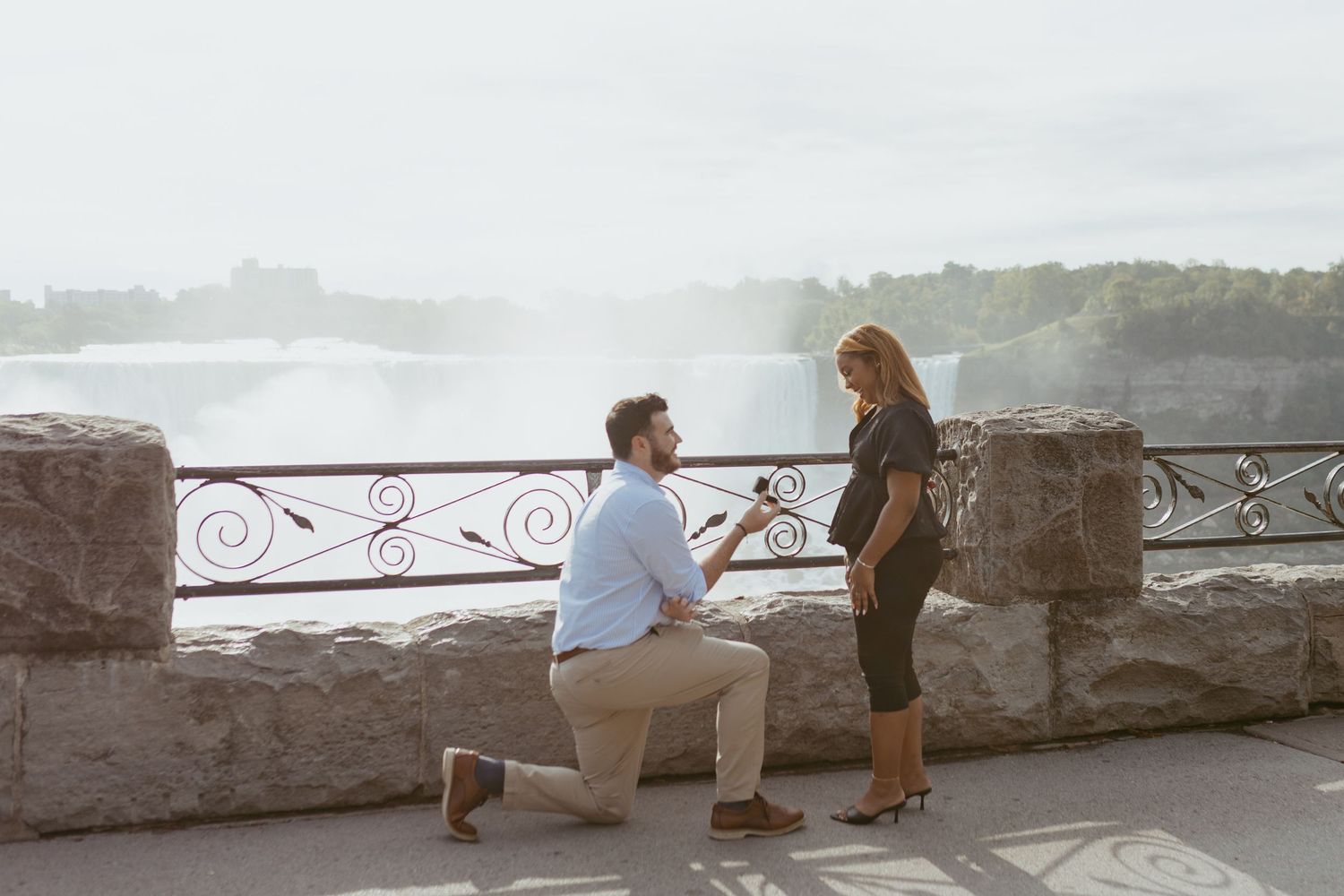 Man proposing to woman on one knee at Niagara Falls overlook with misty waterfall backdrop.