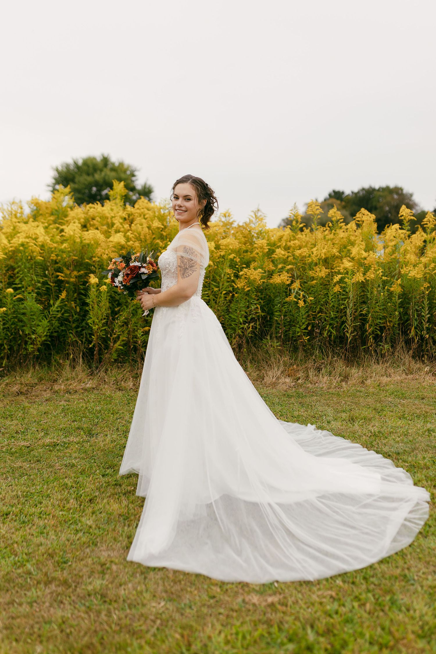 Bride in lace-sleeved wedding gown poses in front of yellow wildflower field.