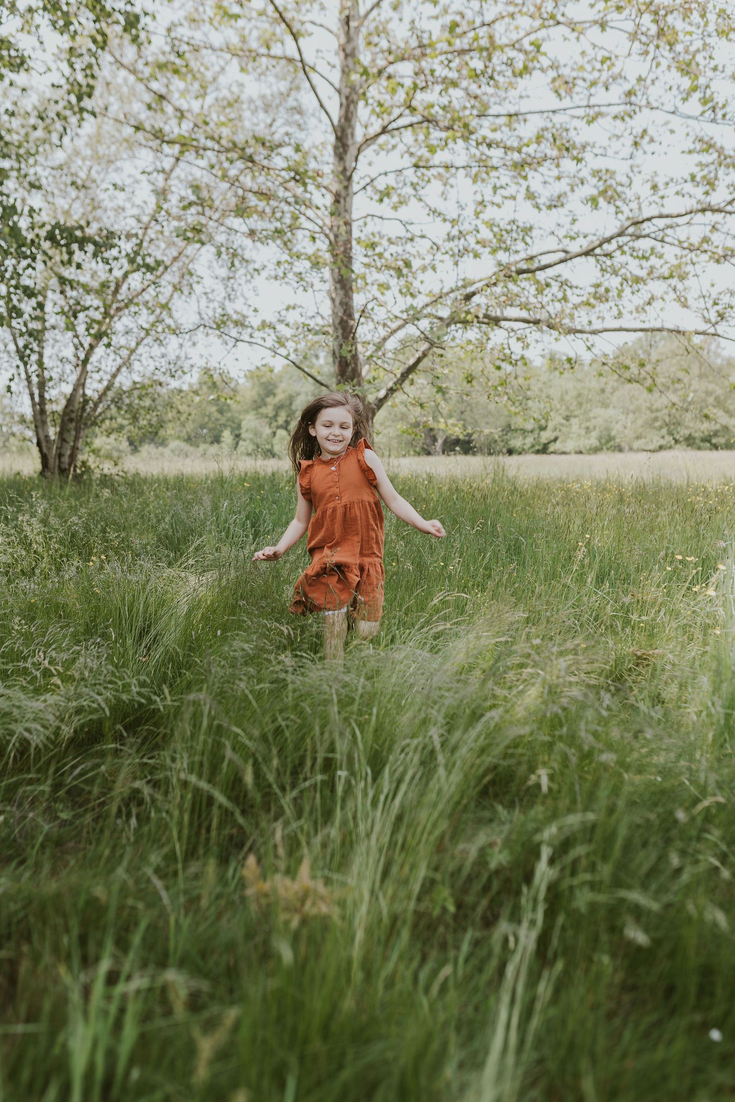 Young person in rust-colored dress running through tall grass field with trees in background.