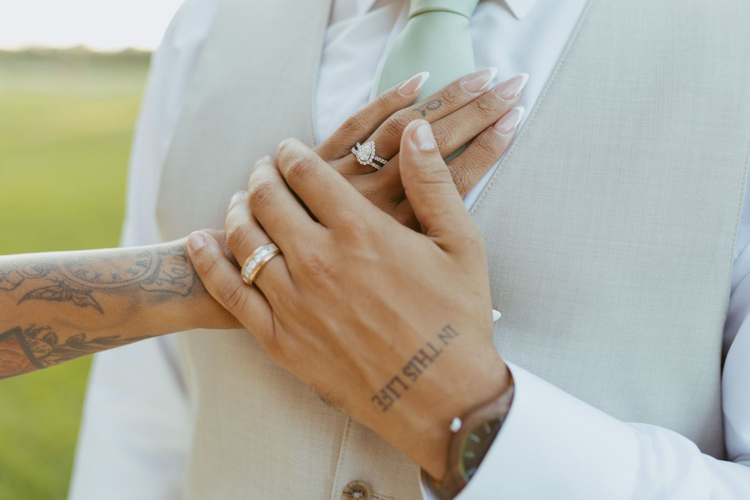 Close-up of wedding couple's hands showing engagement ring and wedding bands, with tattoos visible on wrists.